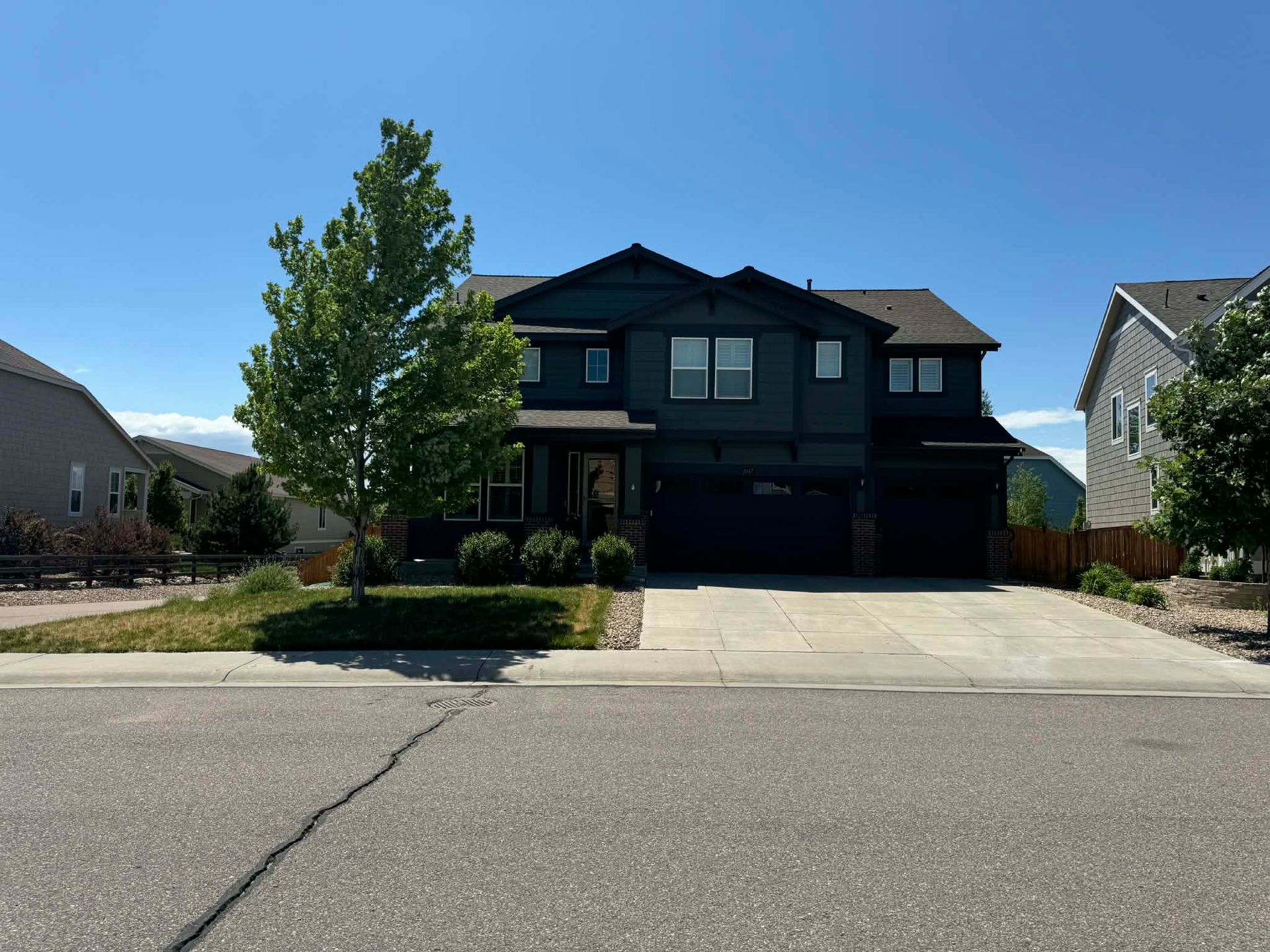 A two-story, dark-colored house with a large green tree in the front yard and a concrete driveway under a clear blue sky.