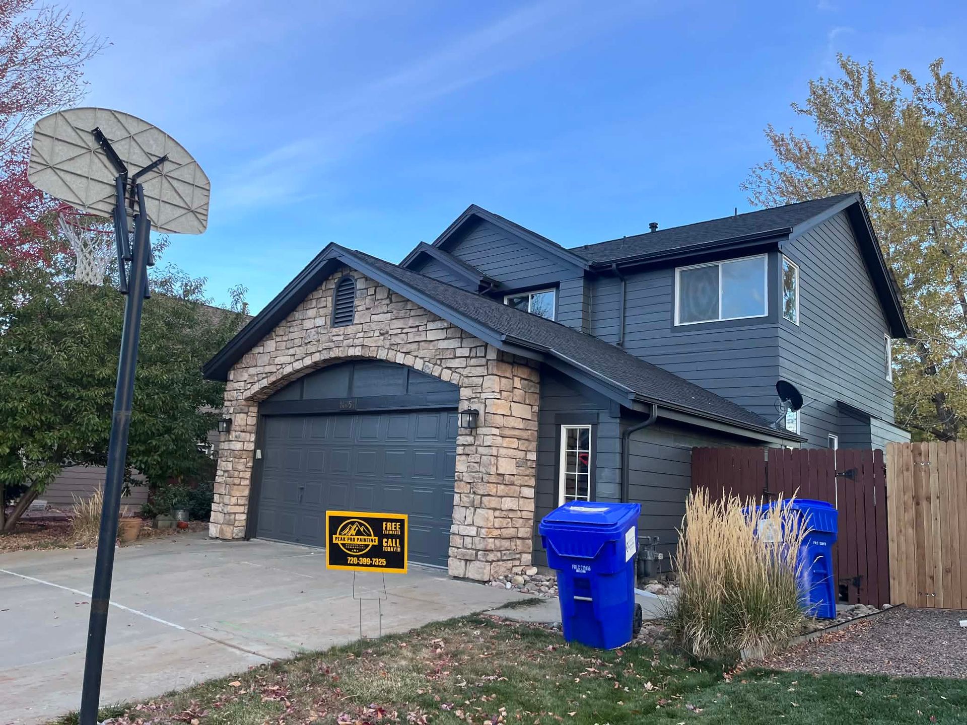 Two-story house with dark gray siding, a stone-front garage, a basketball hoop, and blue recycling bins on the driveway.