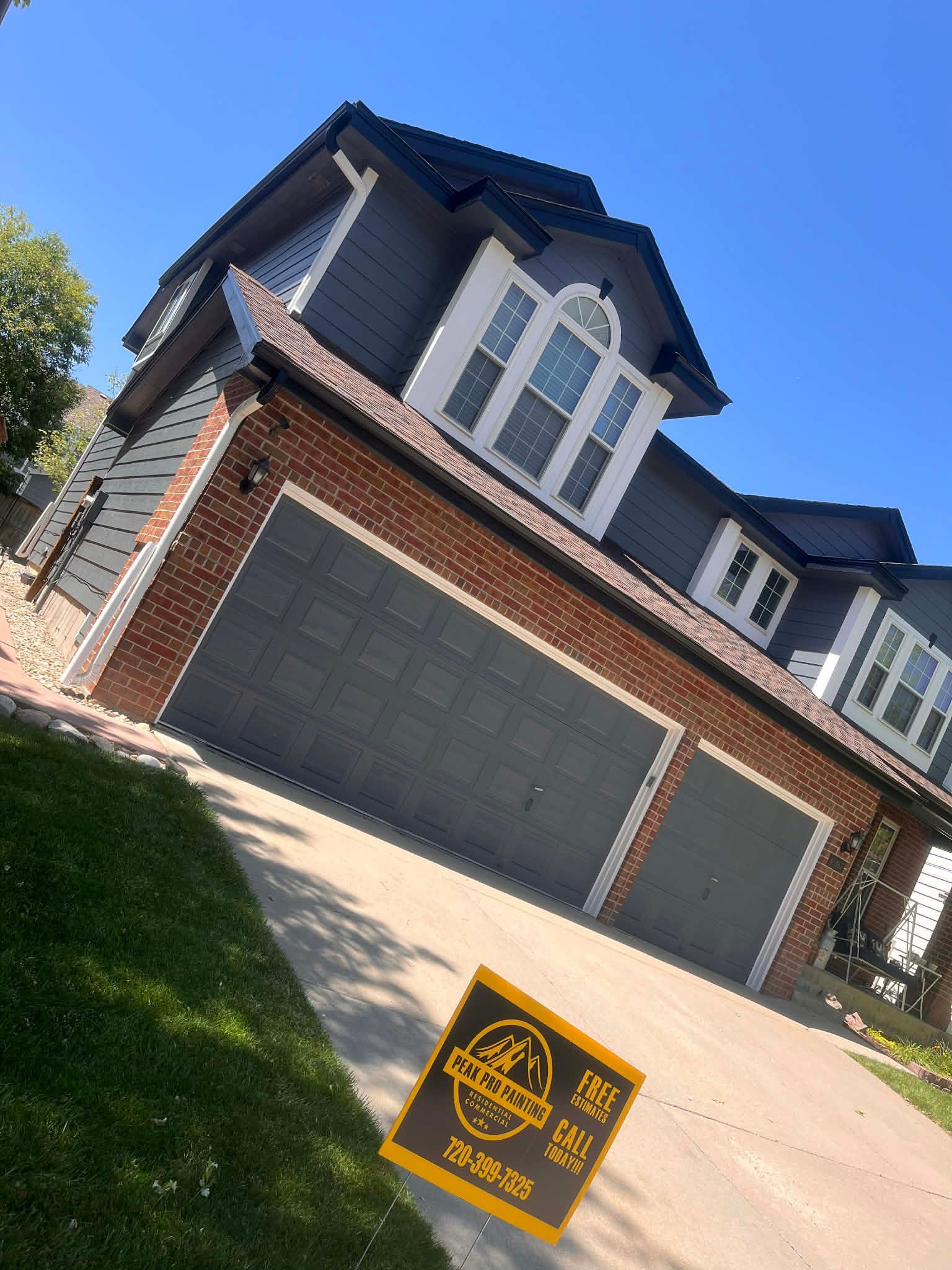 A two-story house with dark gray siding and a red brick garage exterior on a sunny day, with a sign in the driveway.