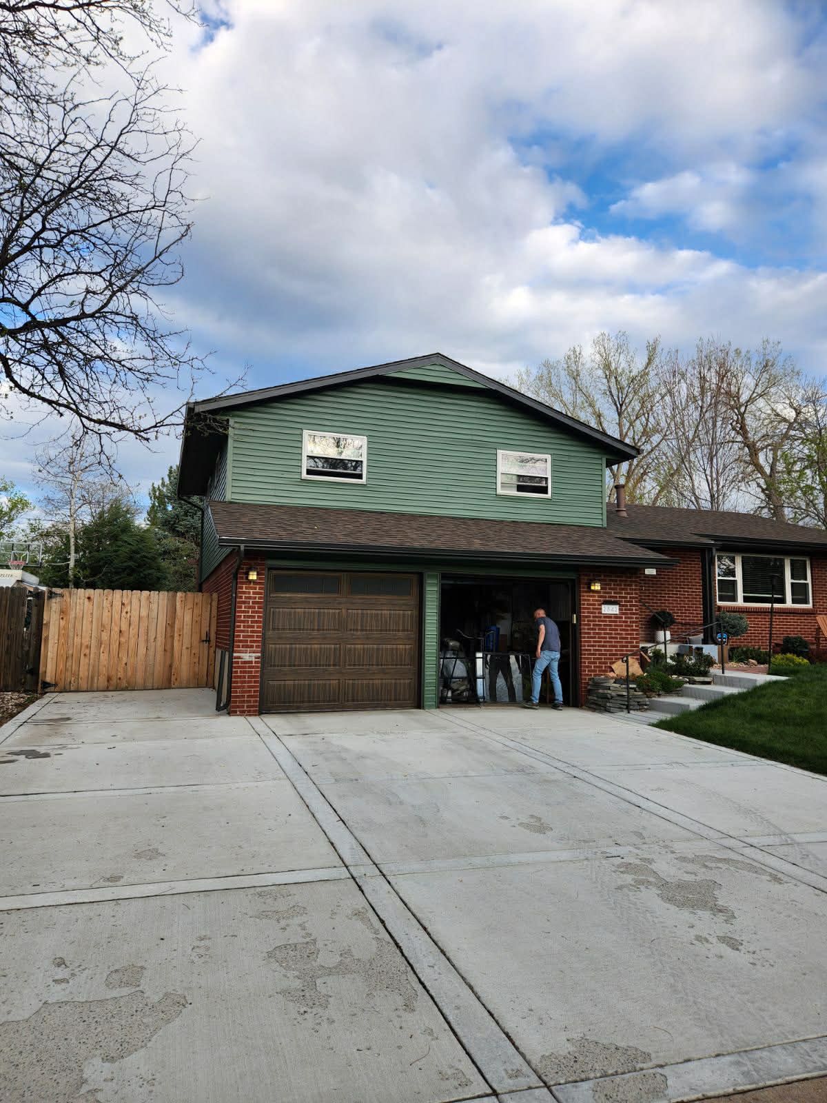 A two-story house with green siding, a dark brick lower level, a two-car garage, and a large concrete driveway.