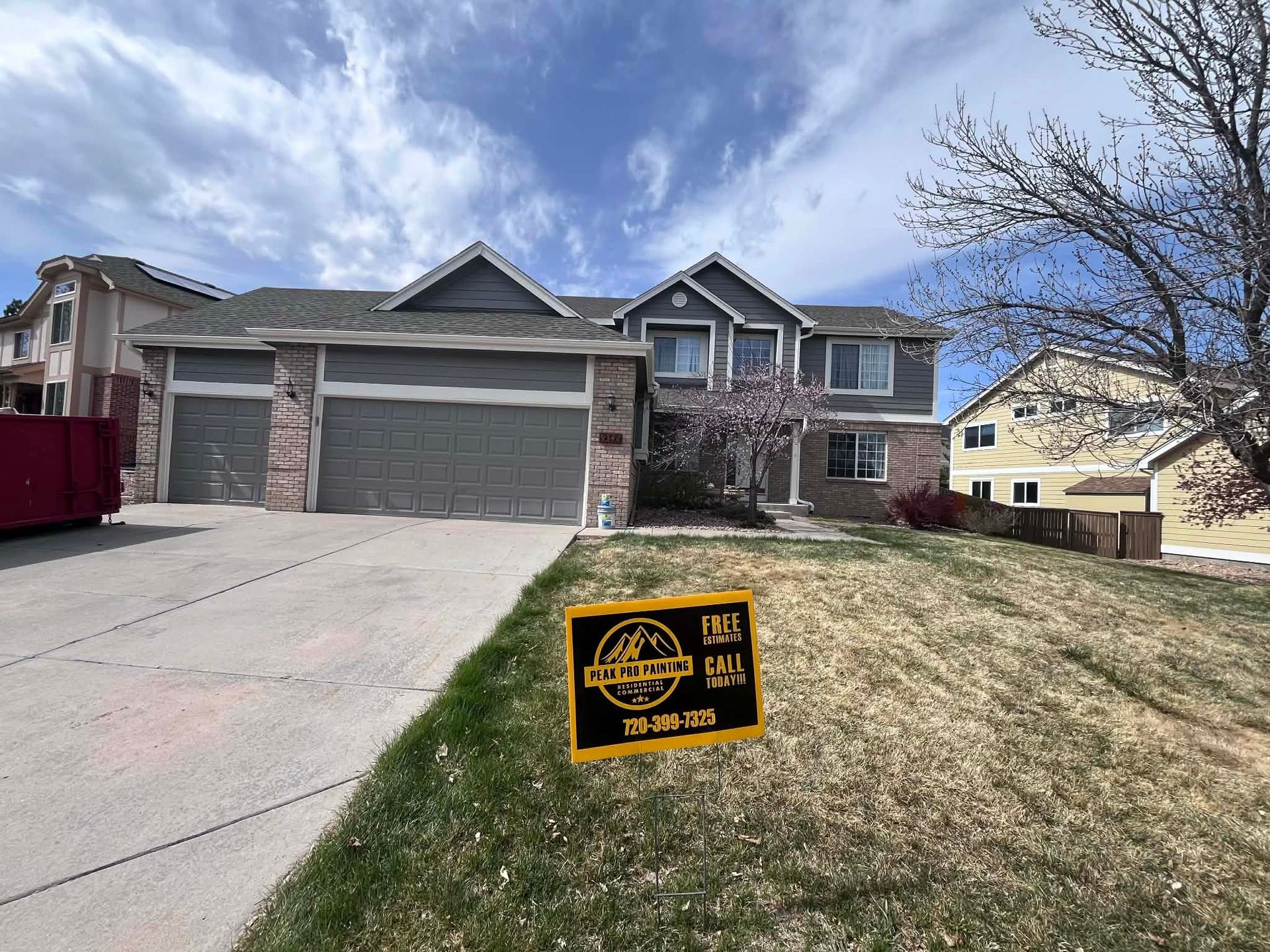 A two-story grey suburban house with a three-car garage and a sign in the front yard under a partly cloudy sky.