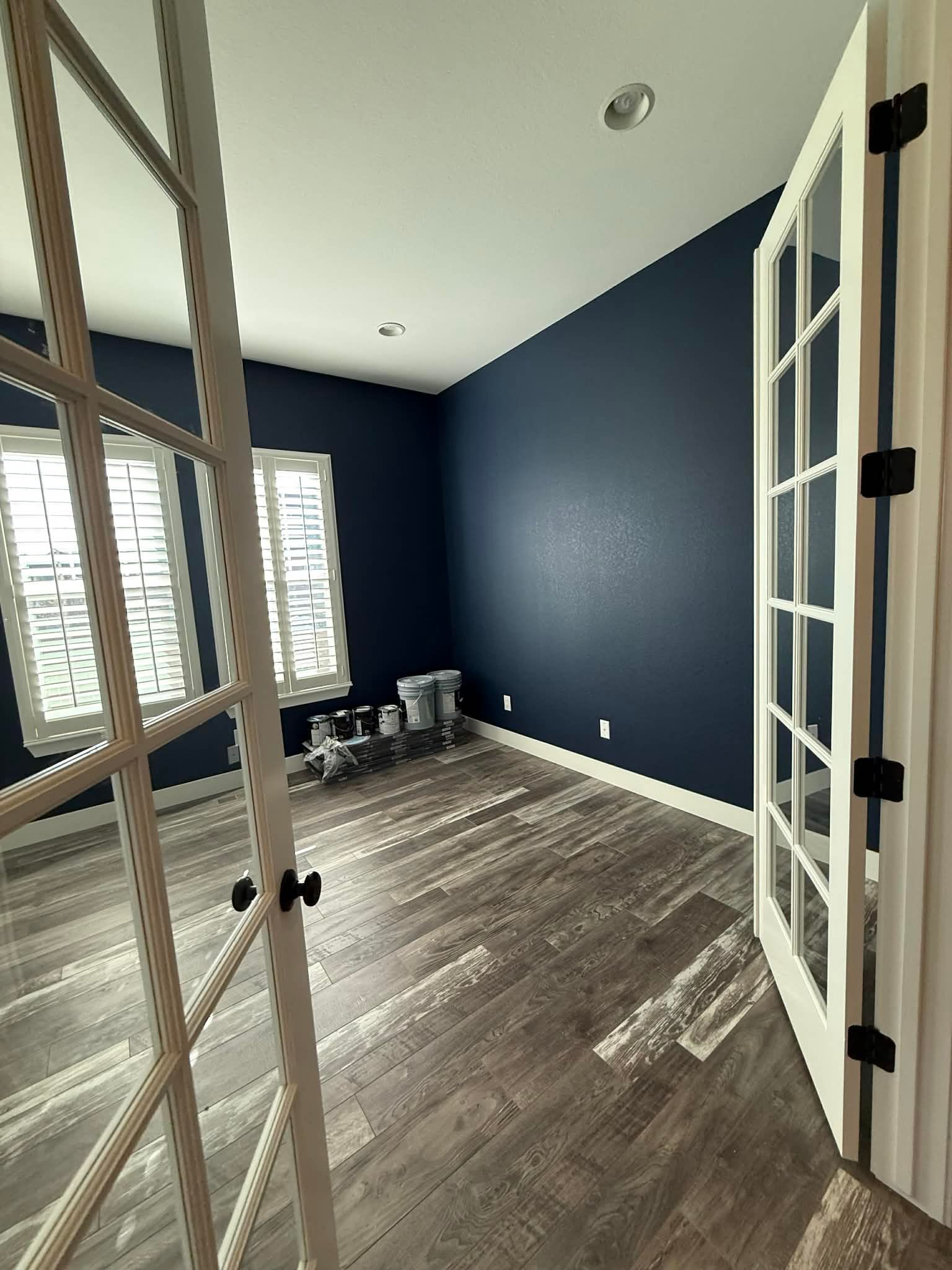An empty room with dark blue walls, light wood-look flooring, two windows with shutters, and white French doors.