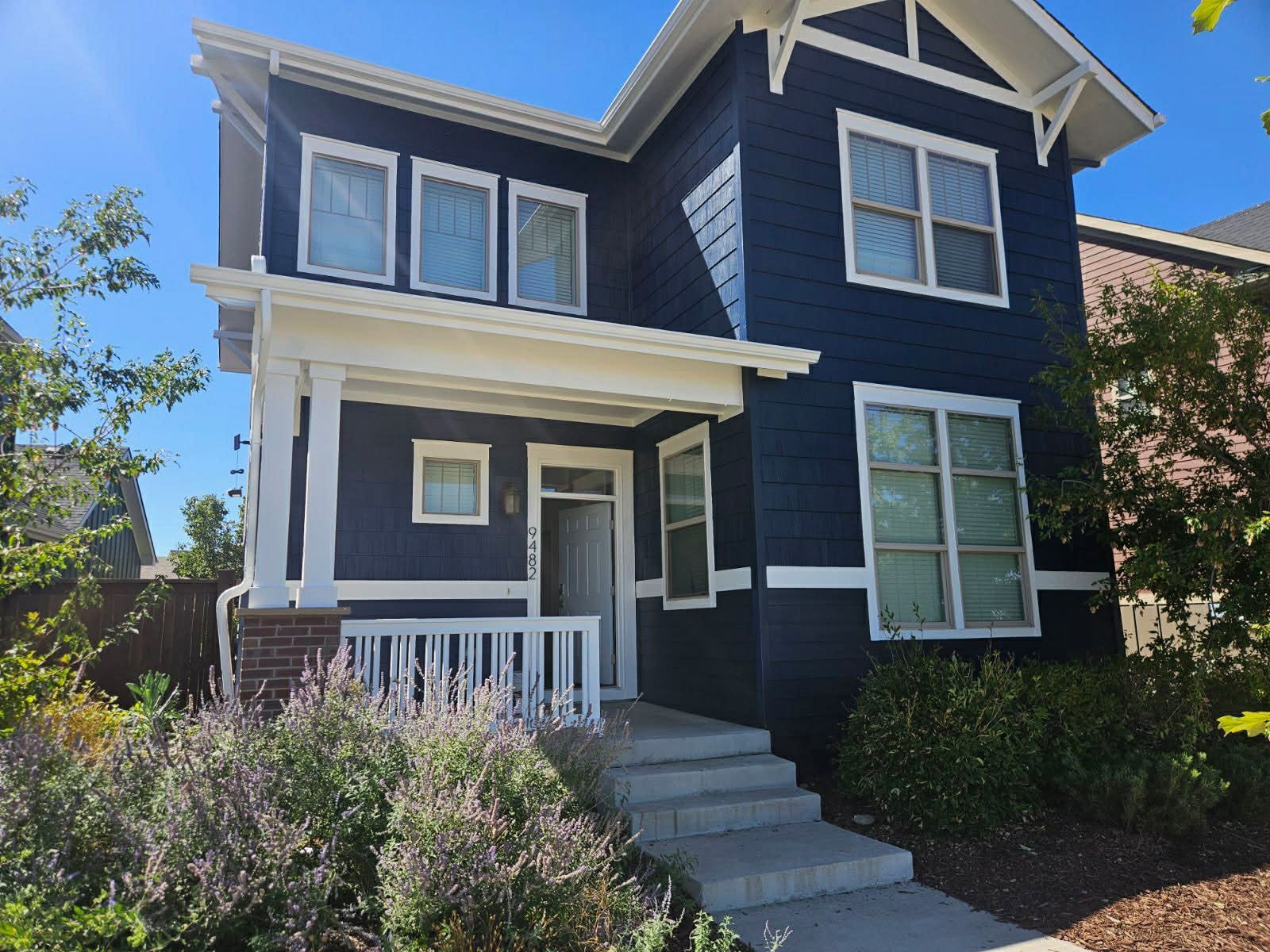A two-story dark blue suburban house with white trim, a front porch, and steps leading to the entrance on a sunny day.
