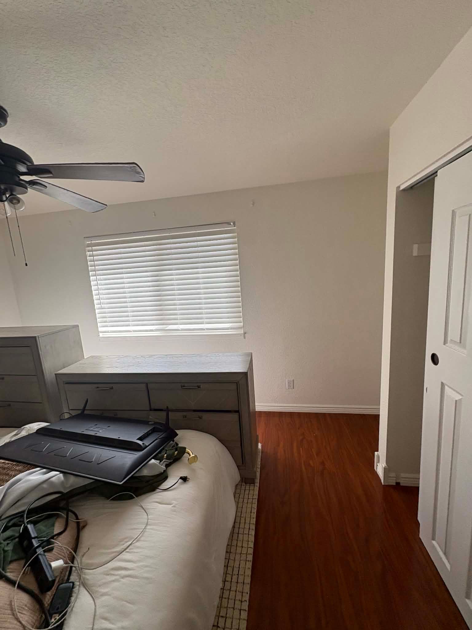 A bedroom featuring a dresser with a television on top, a bed in the foreground, a window with blinds, and wood floors.