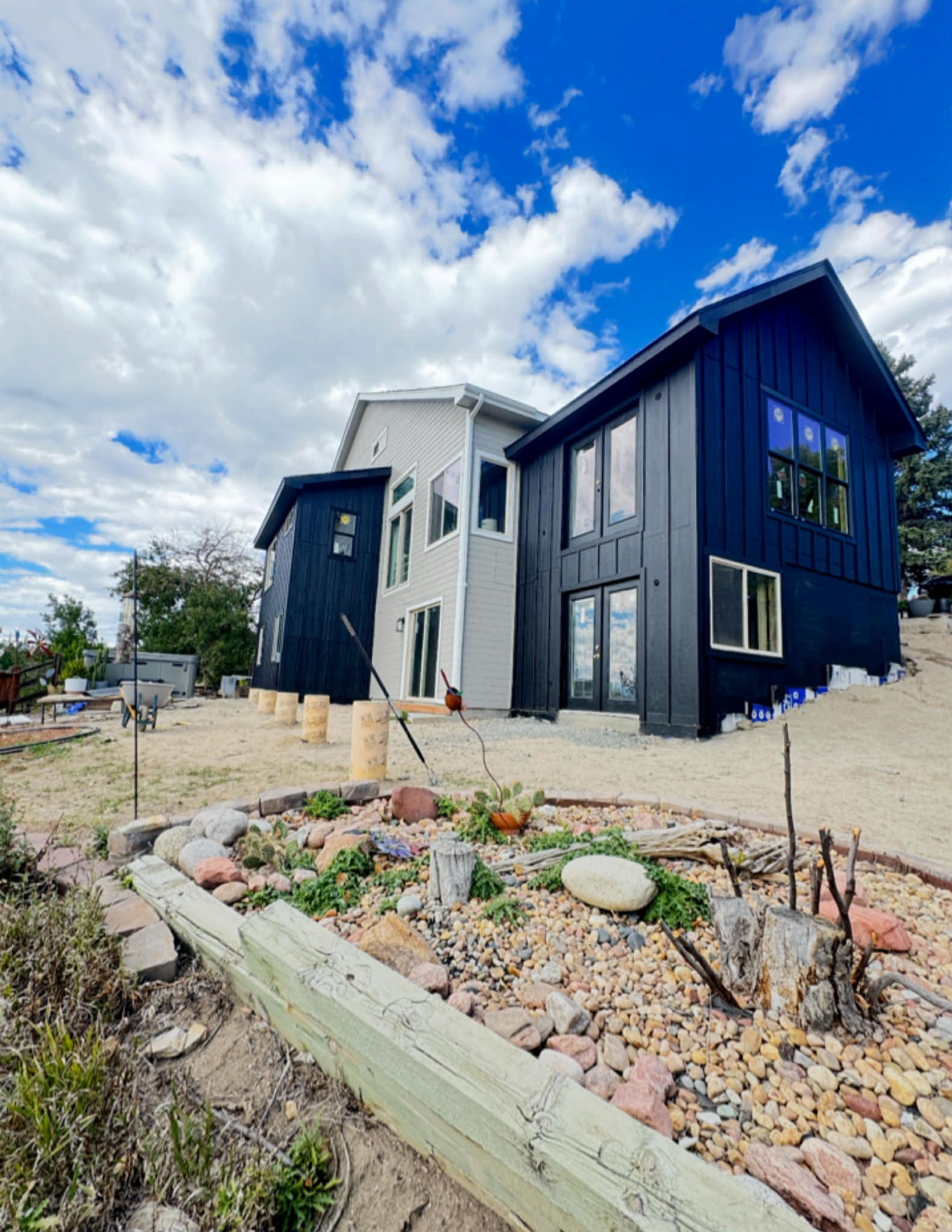 A modern two-story home with a mix of black vertical siding and white walls, set on a gravel lot with a rock garden.