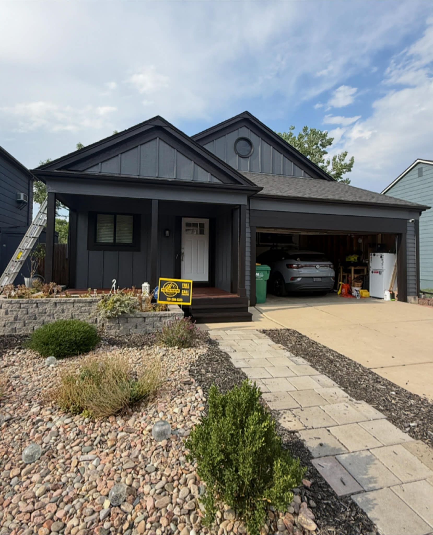 Dark gray house with a peaked roof, covered front porch, a parked car inside the garage, and a rock-landscaped front yard.