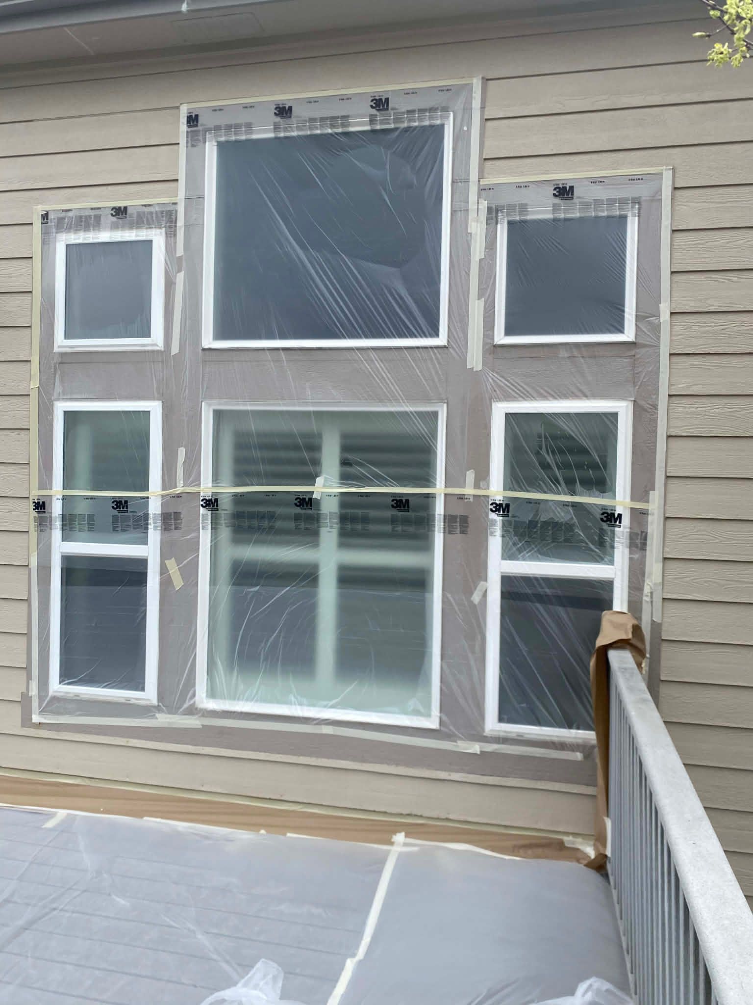 A large, multi-pane window assembly covered in protective plastic film on the exterior of a light brown sided house.