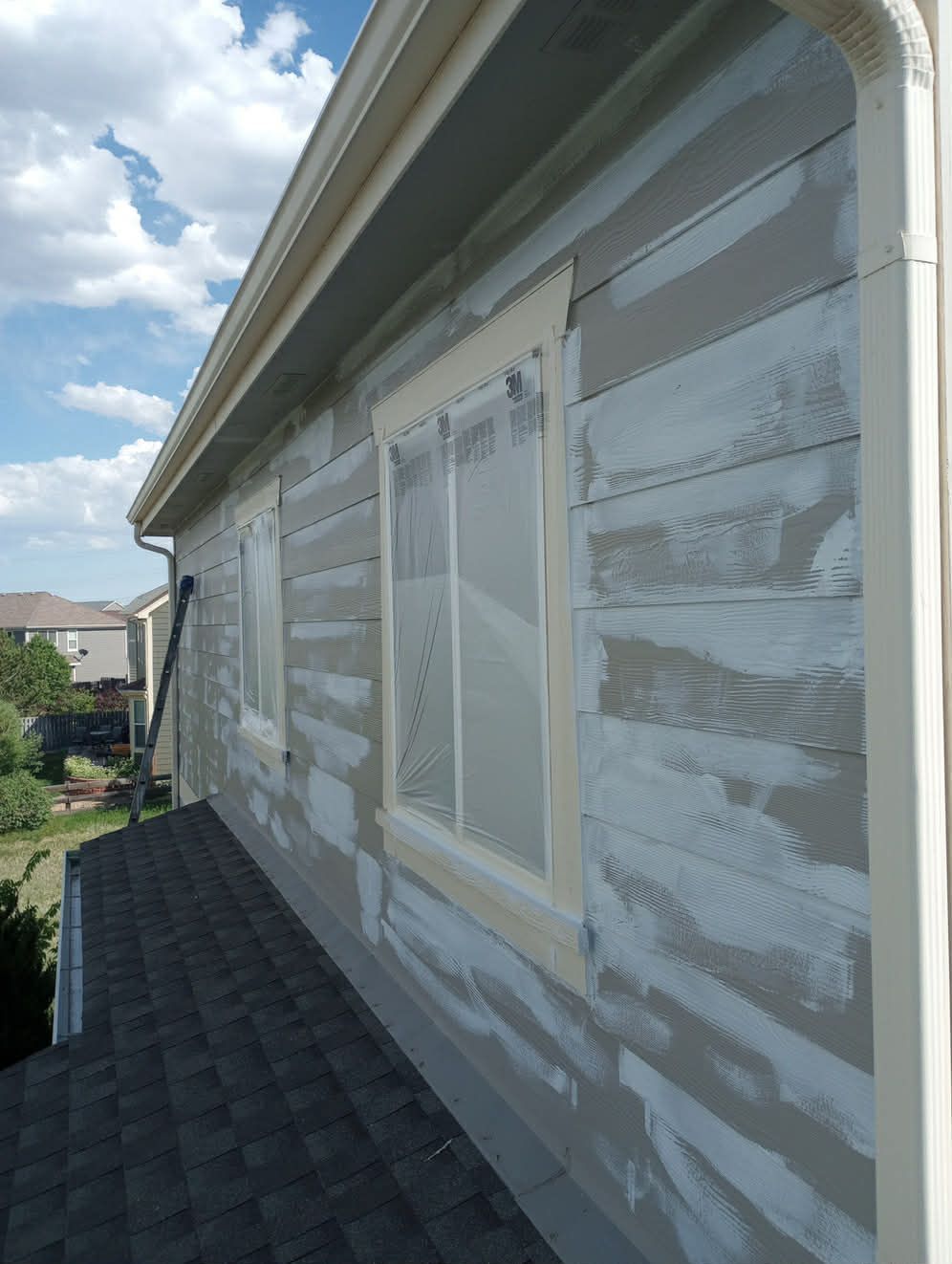 Exterior side of a house undergoing repairs with patchy primer on the siding and taped-off windows above a shingled roof.