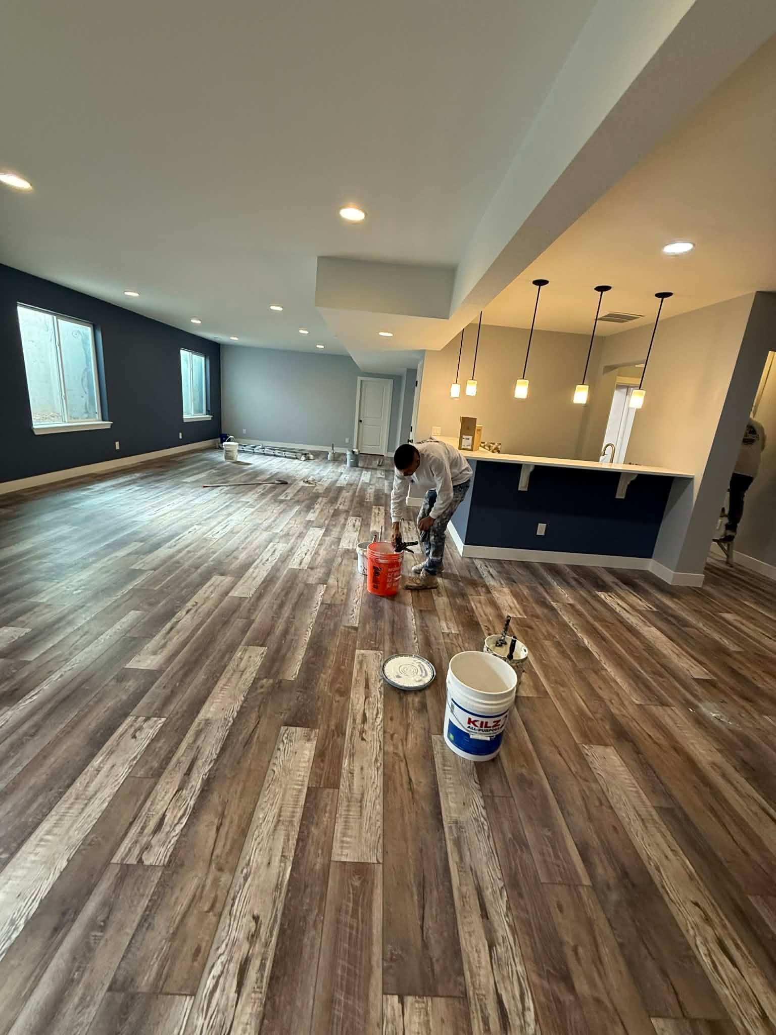 A person works in a large, modern finished basement with wood-look vinyl flooring, a home bar, and dark grey accent walls.
