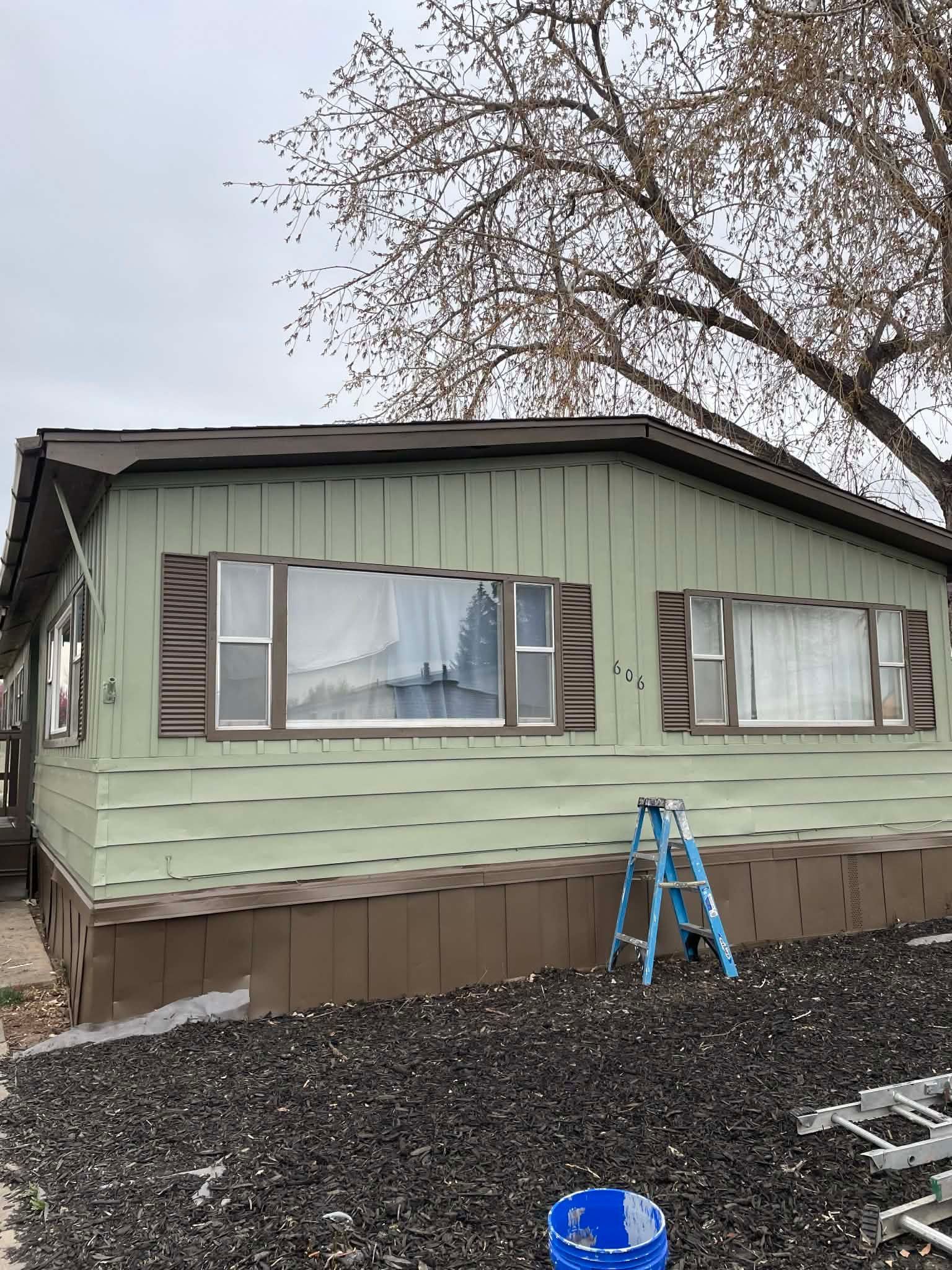 A light green mobile home with brown trim and shutters, with a blue stepladder in front and gravel on the ground.