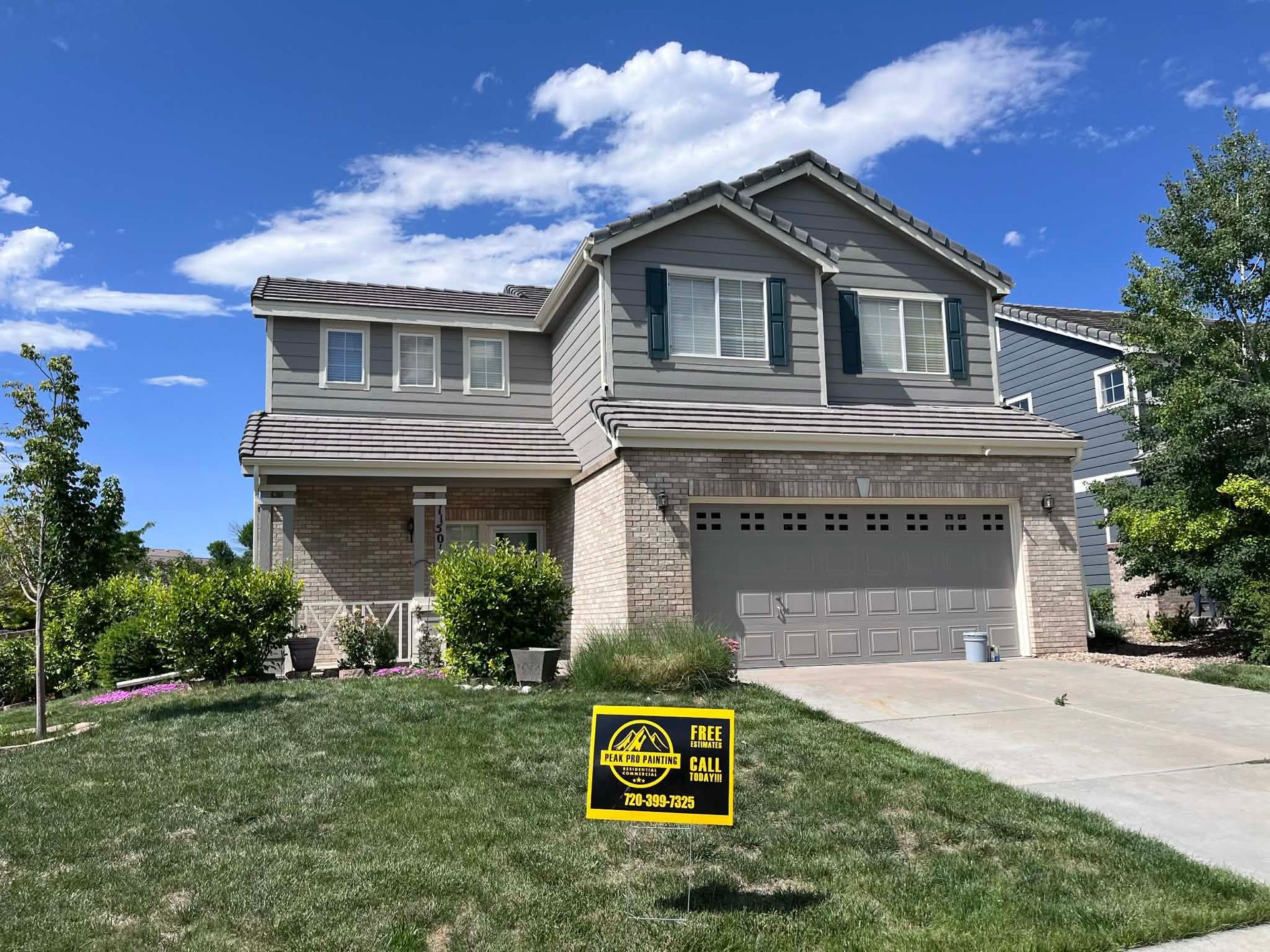 A two-story suburban house with beige brick, gray siding, a double garage, and a yellow real estate sign on the lawn.