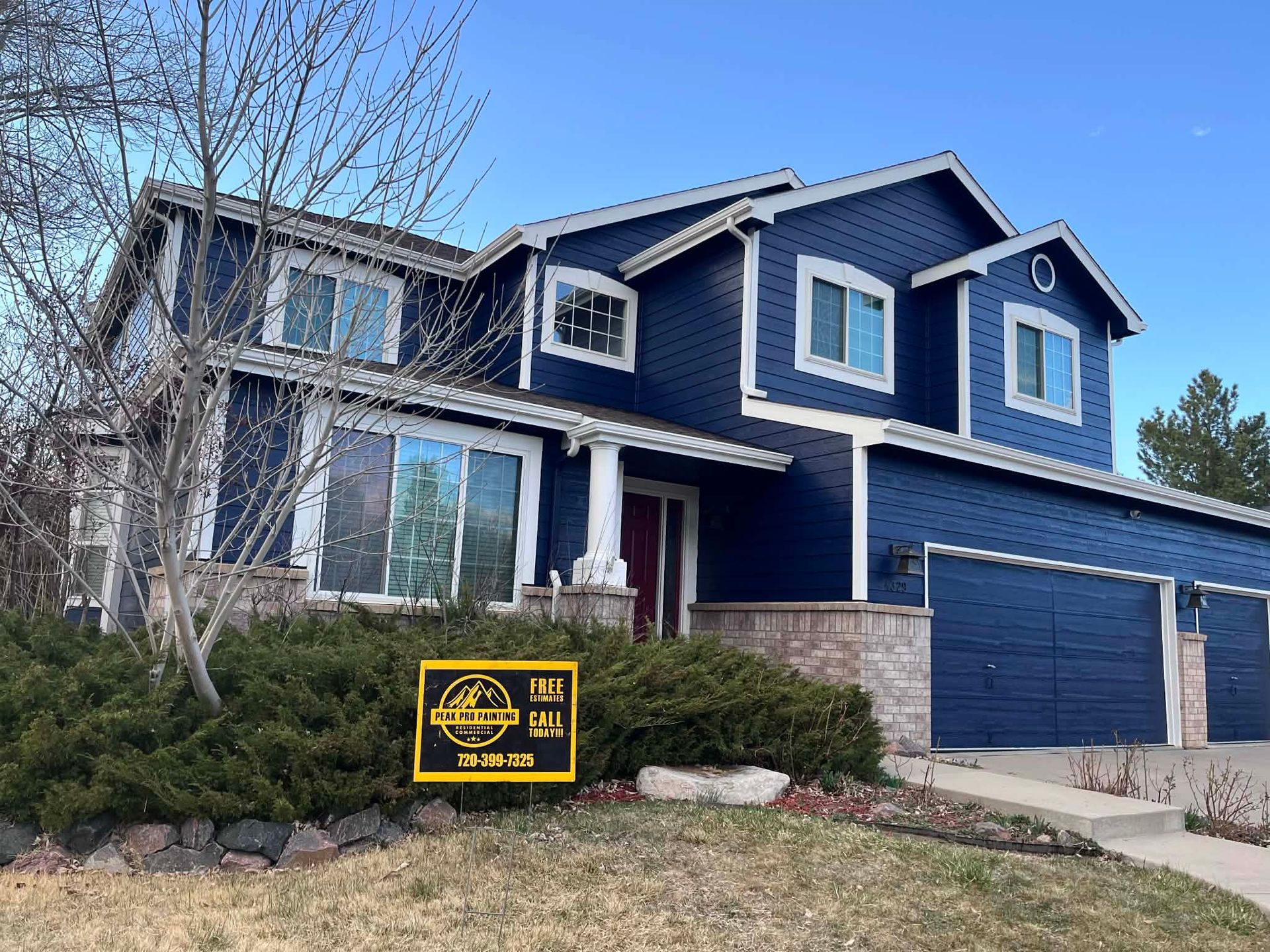 A two-story blue house with a three-car garage and a small sign in the front landscaping.