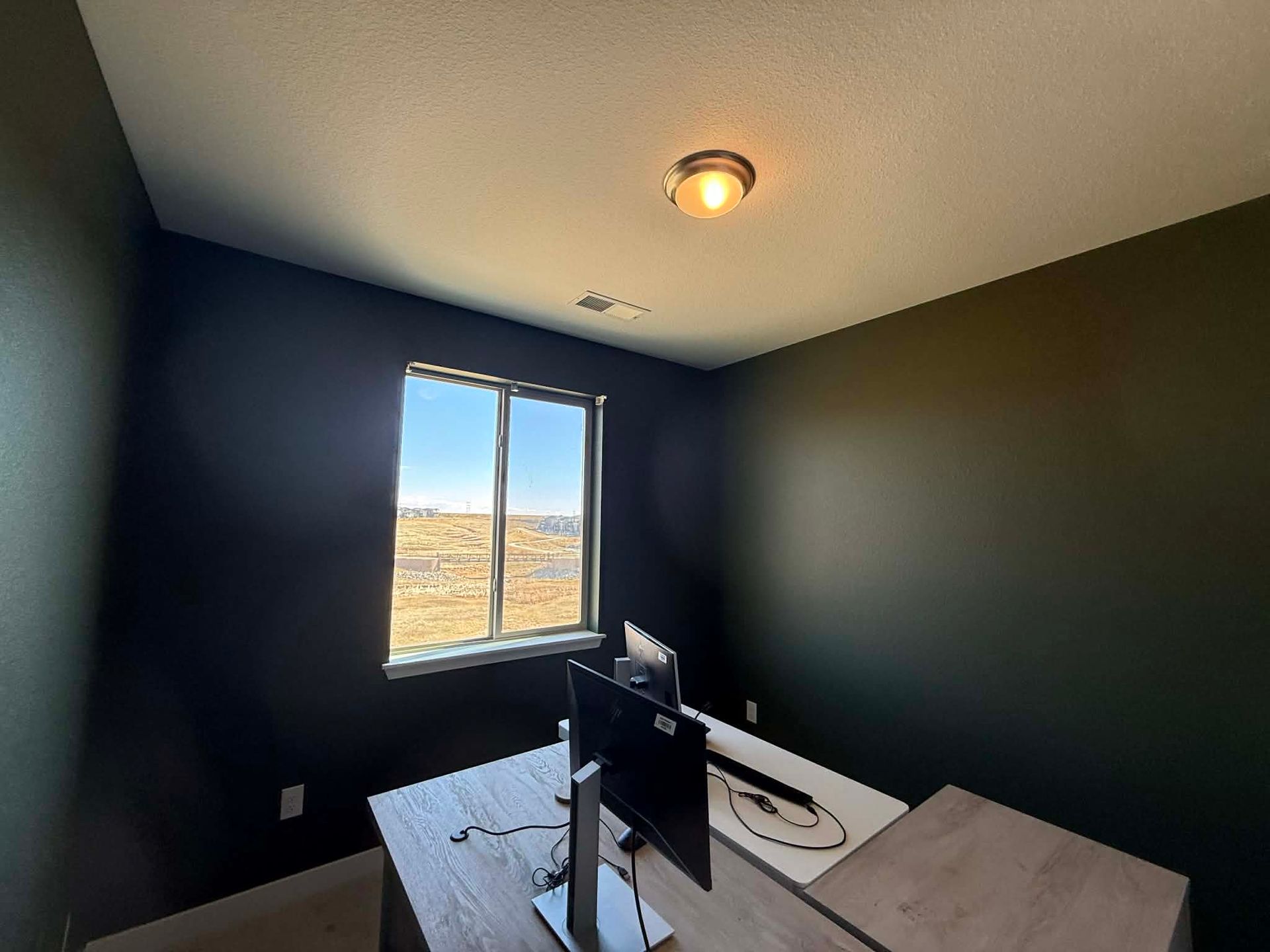 A home office with dark charcoal walls, a wooden desk with a computer monitor, and a single window overlooking a field.