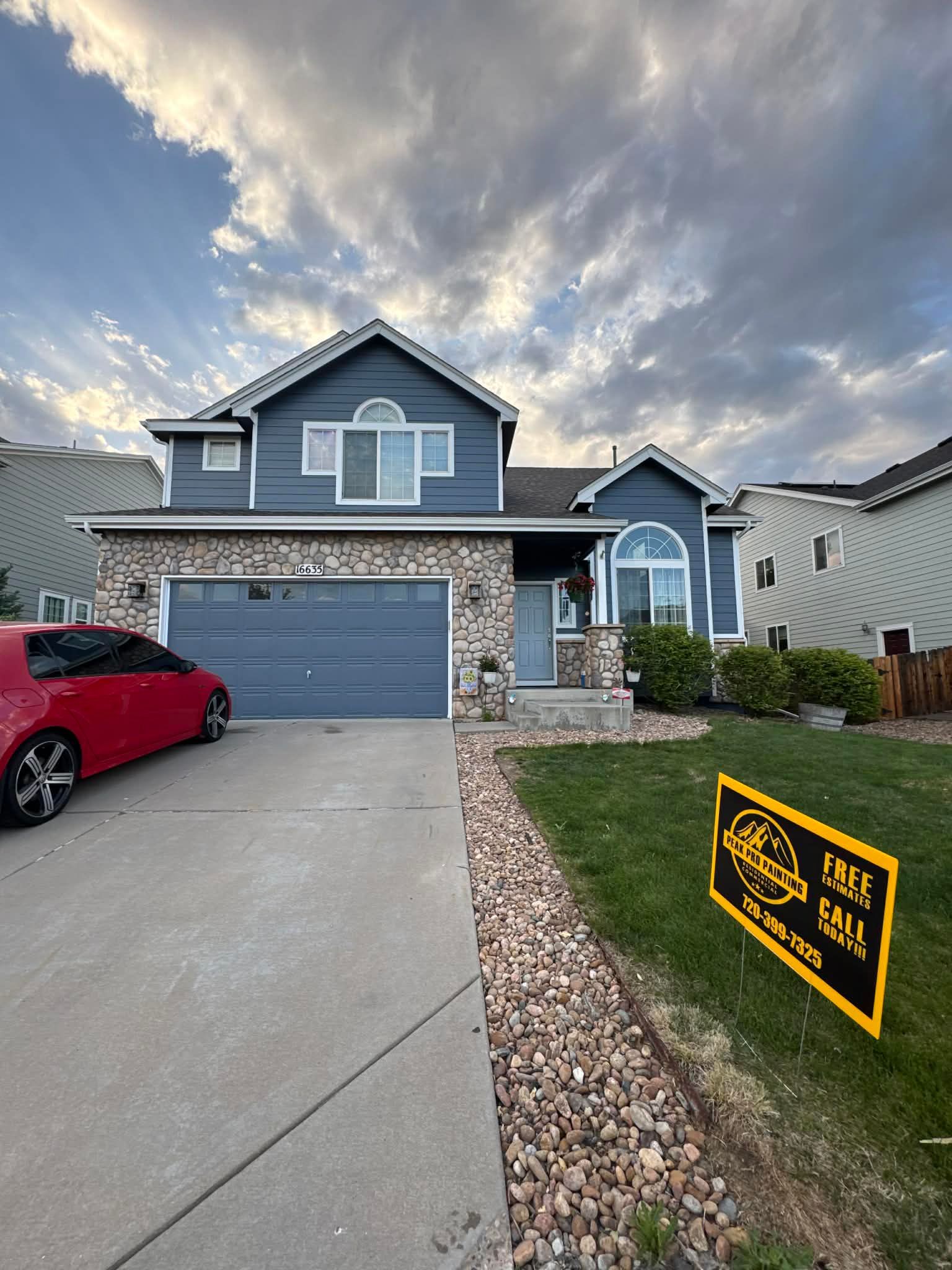 A blue, two-story house with a stone facade, a gray garage door, and a red car parked in the driveway at sunset.