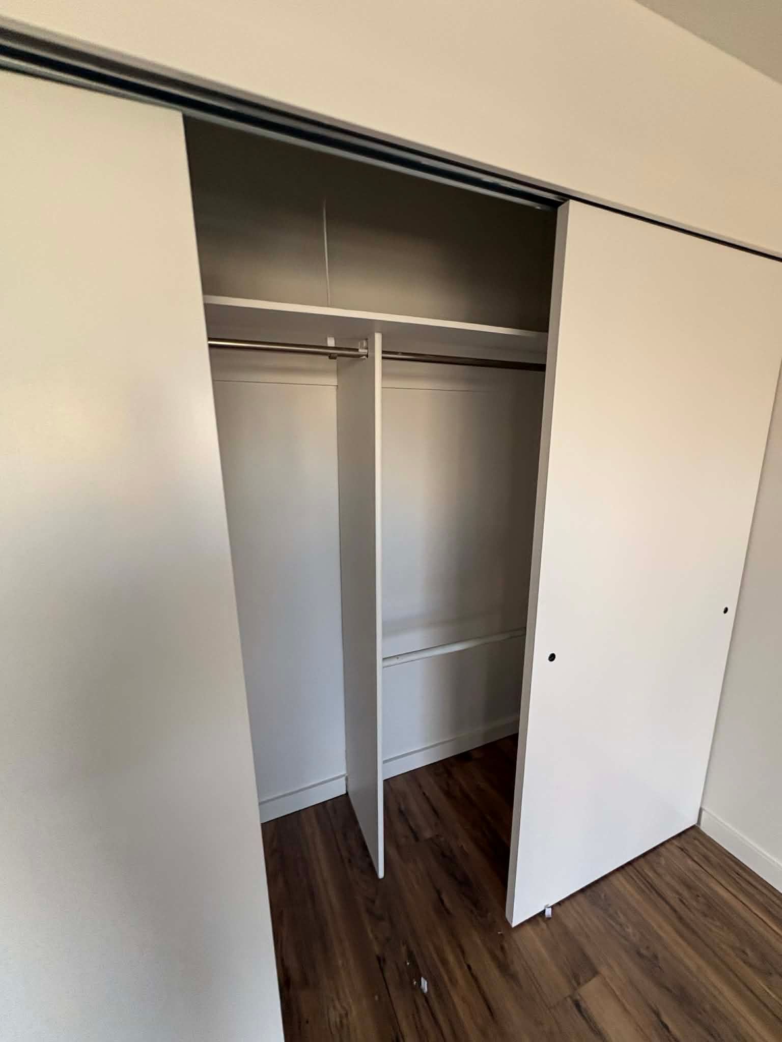 A white, sliding-door closet in a room with wood-look flooring, showing an open interior with shelves and a hanging rod.