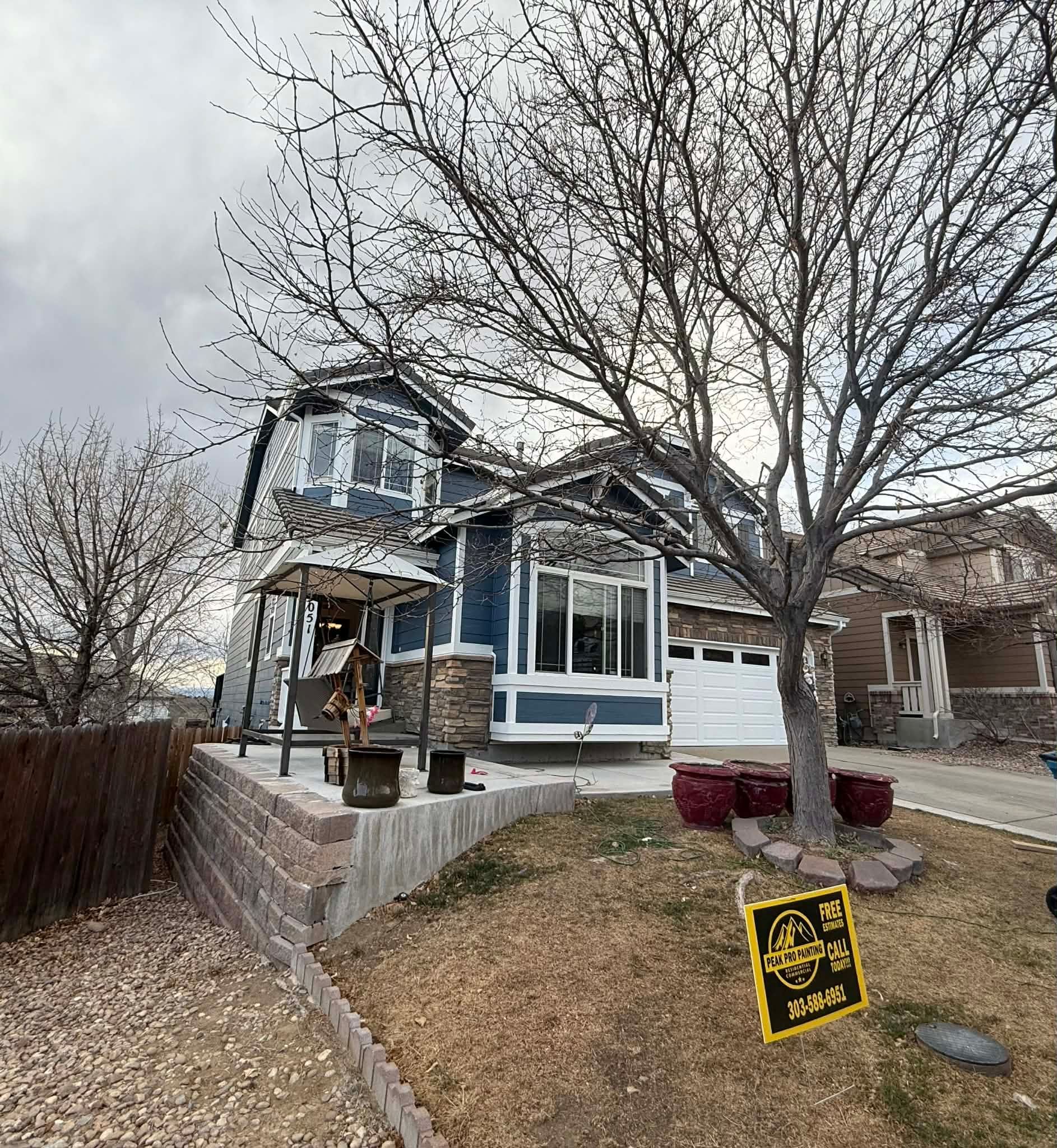 A two-story blue house with stone accents, a landscaped front yard with a tree, and a yellow sign in the grass.