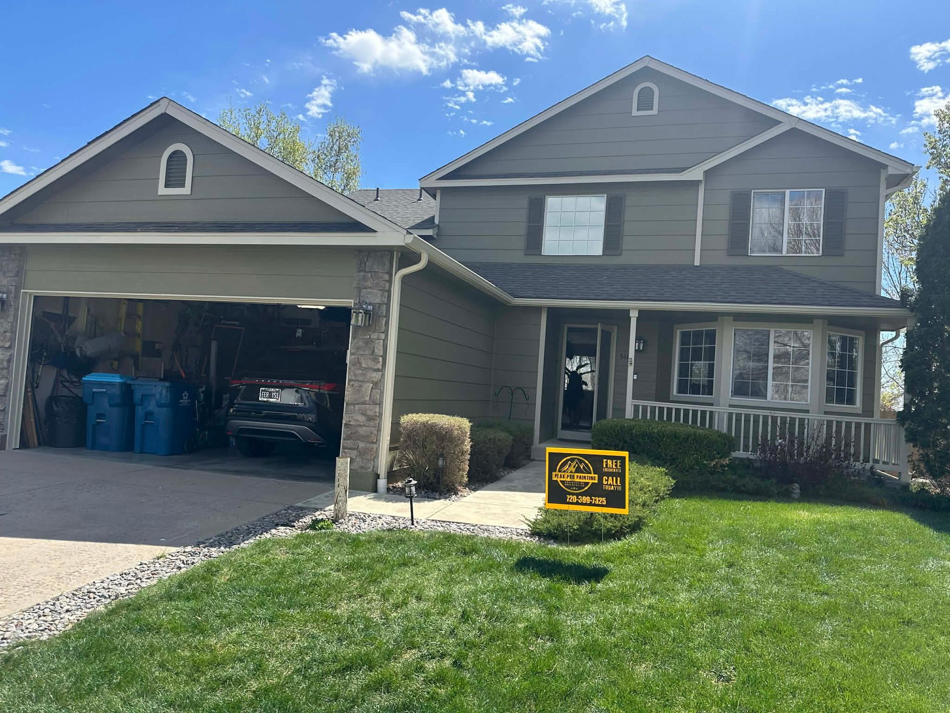 A two-story olive-green house with an open garage, a front porch, and a yellow real estate sign on the lawn.