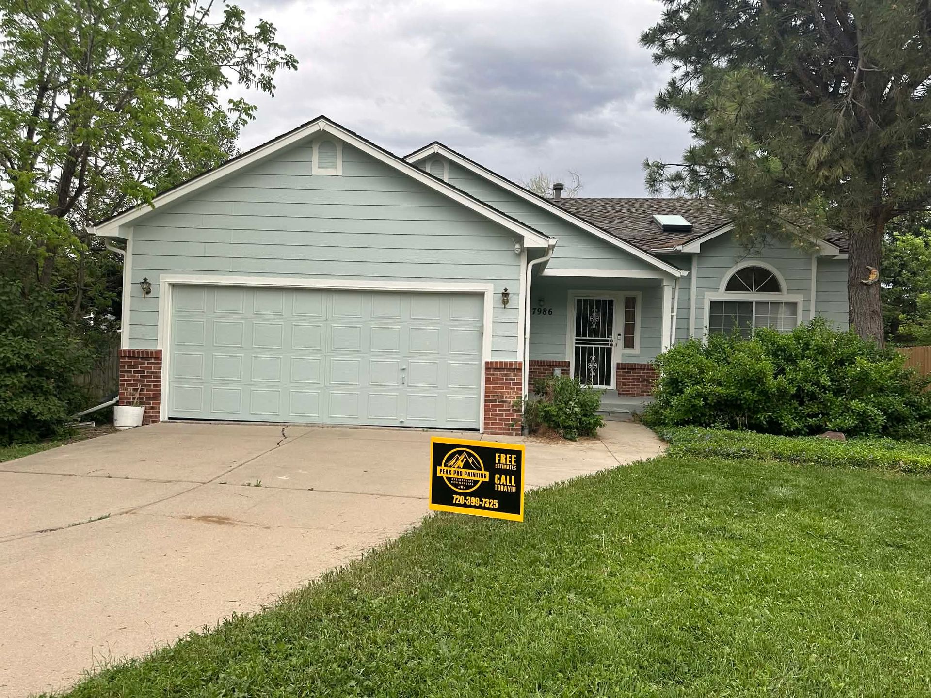 Light green single-story house with a white garage door, brick accents, and a front lawn under a cloudy sky.