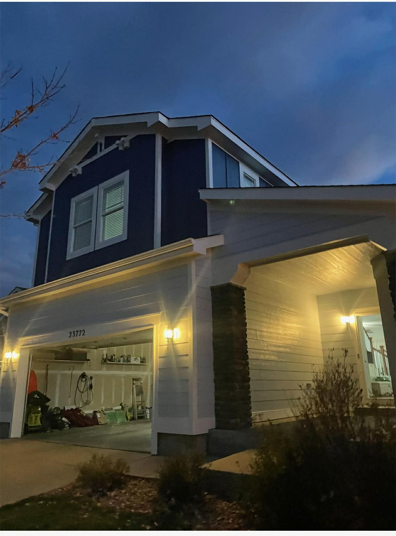 A two-story blue and white house at dusk with an open garage, exterior lights illuminated, and a stone-accented porch.