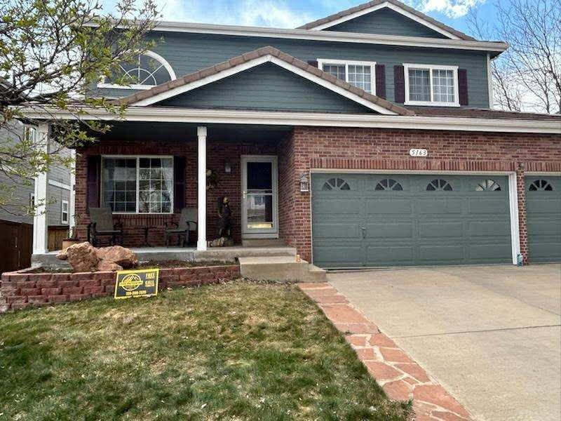 A two-story house with dark green siding, red brick accents, and a large three-car garage, featuring a front porch.
