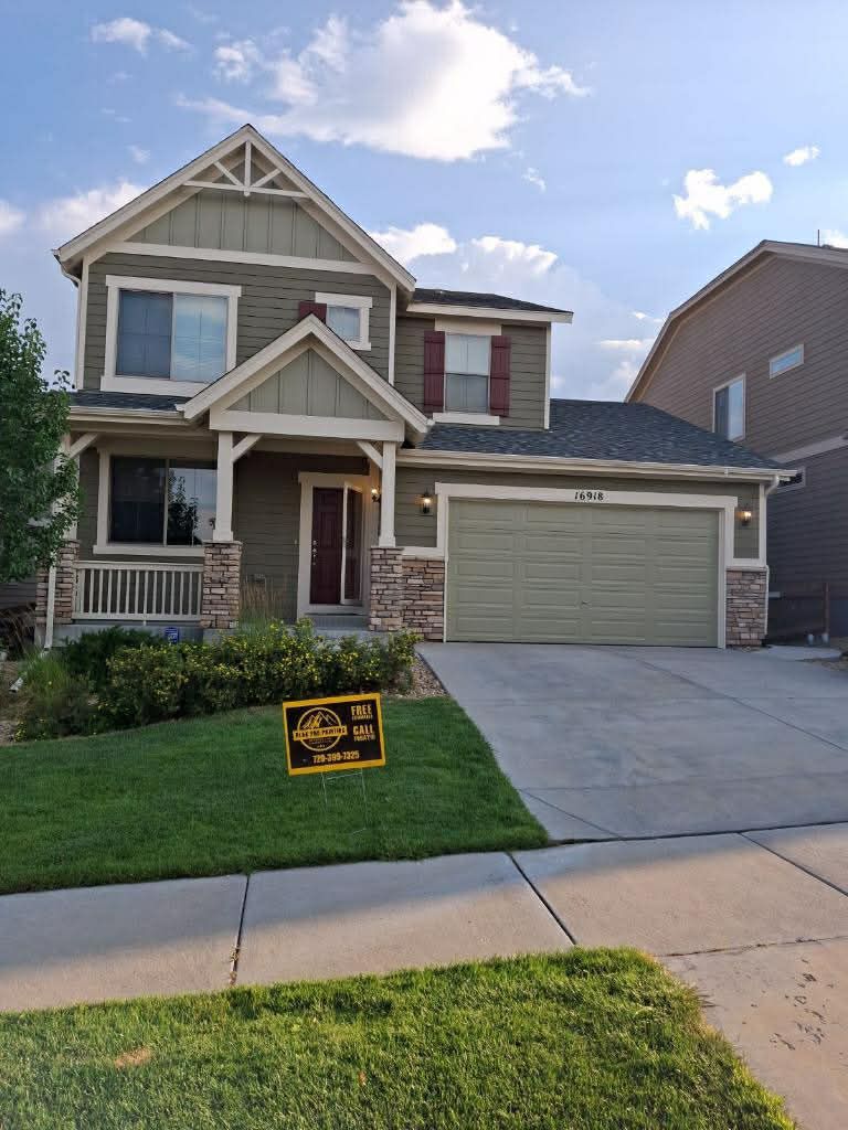 A two-story green suburban house with a two-car garage, stone accents, and a yard sign, seen from the street.