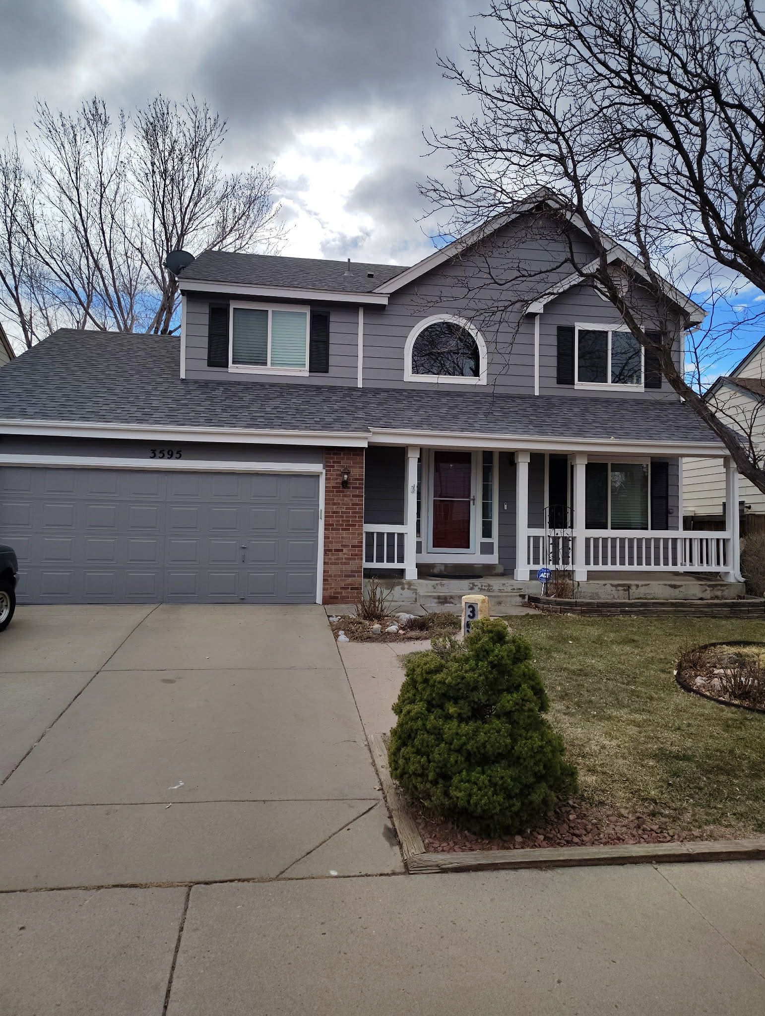 A two-story grey suburban house with a two-car garage, a front porch, and a concrete driveway under a cloudy sky.