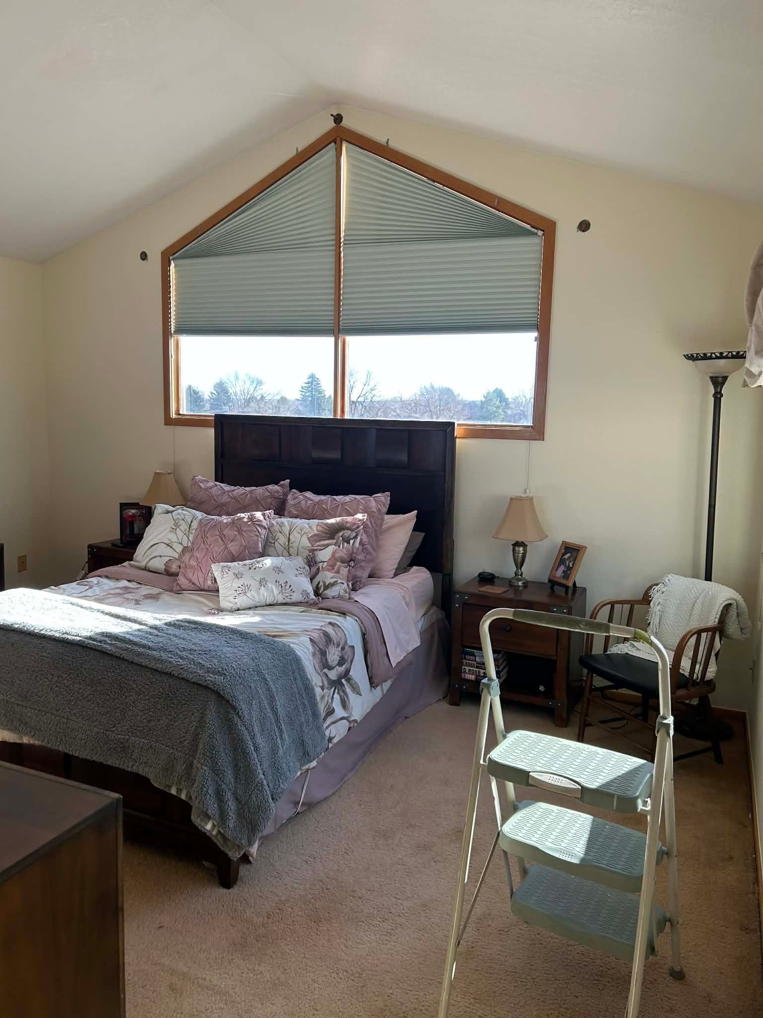 A bedroom with a large triangular window, a dark wood bed with floral bedding, nightstands, and a folding step ladder.