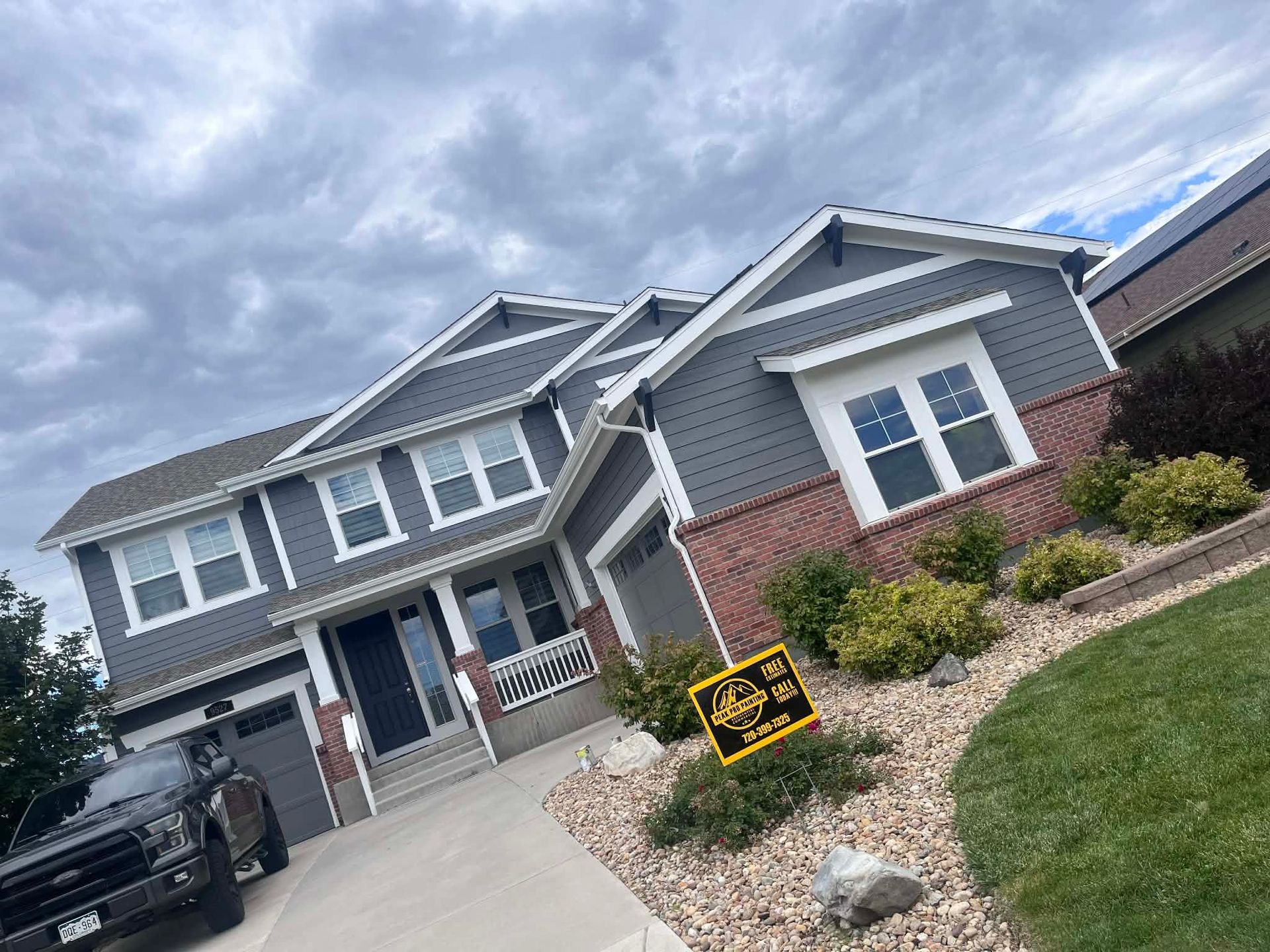 A two-story grey house with brick accents and a driveway featuring a parked truck under a cloudy sky.