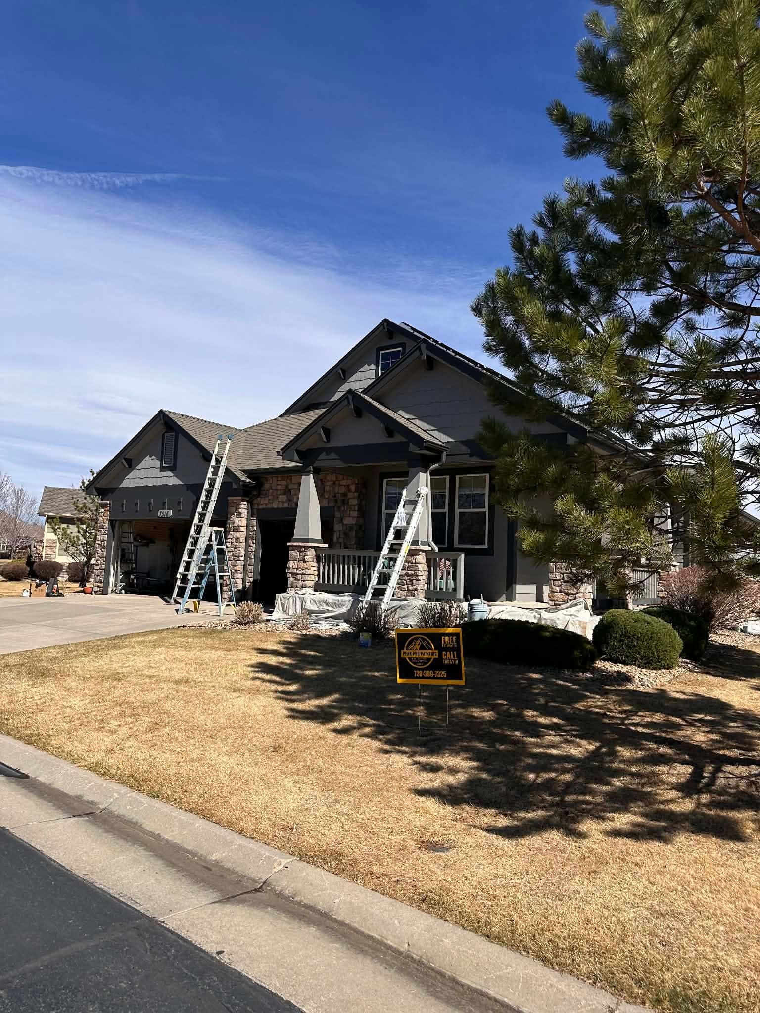 A grey house with stone accents and two ladders leaning against the front exterior on a sunny day.