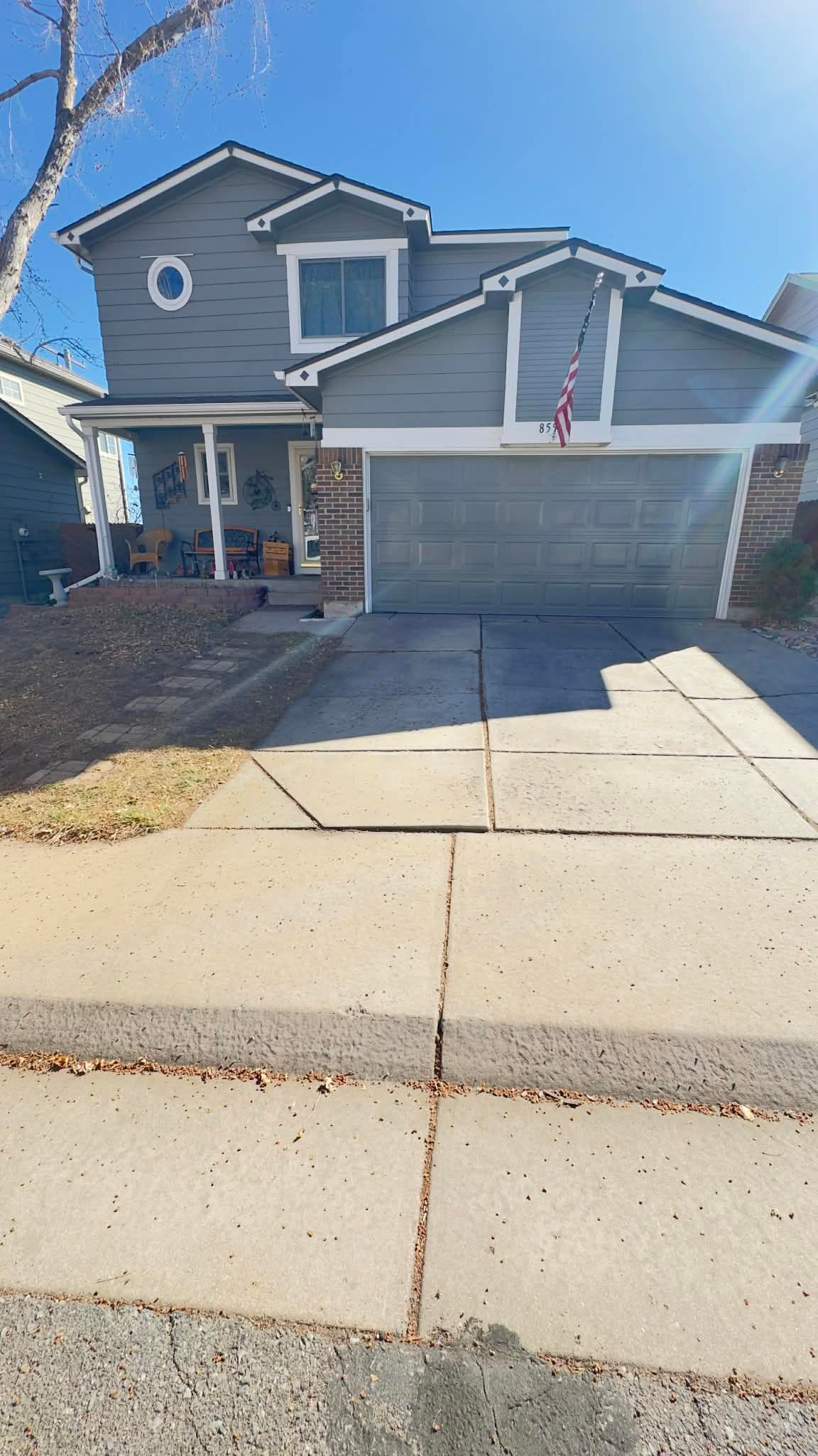 A two-story grey suburban house with a paved driveway, front porch, and garage under a clear blue sky.