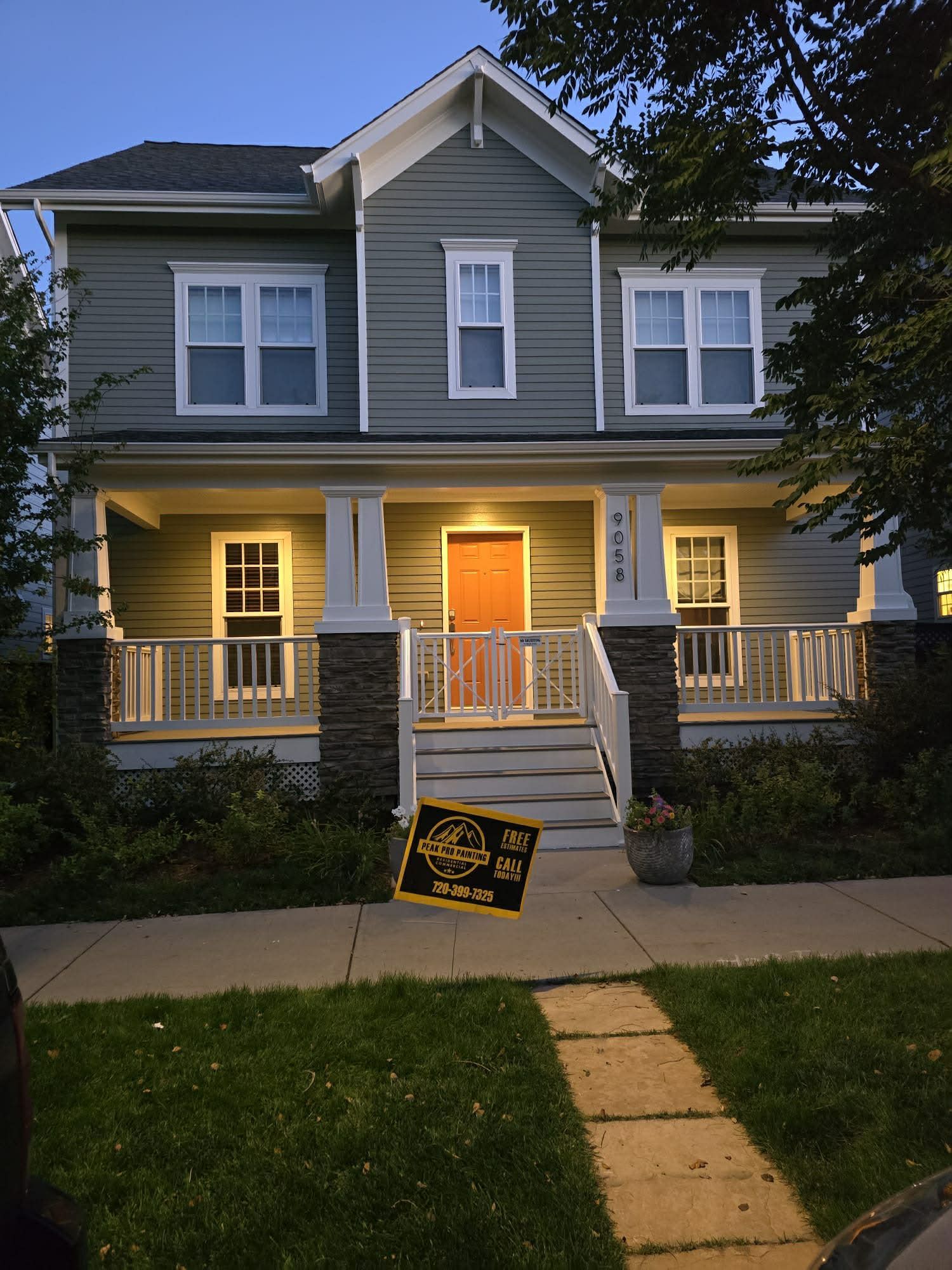 A two-story green-sided house with a front porch, stone pillars, white railings, and an orange door at dusk.