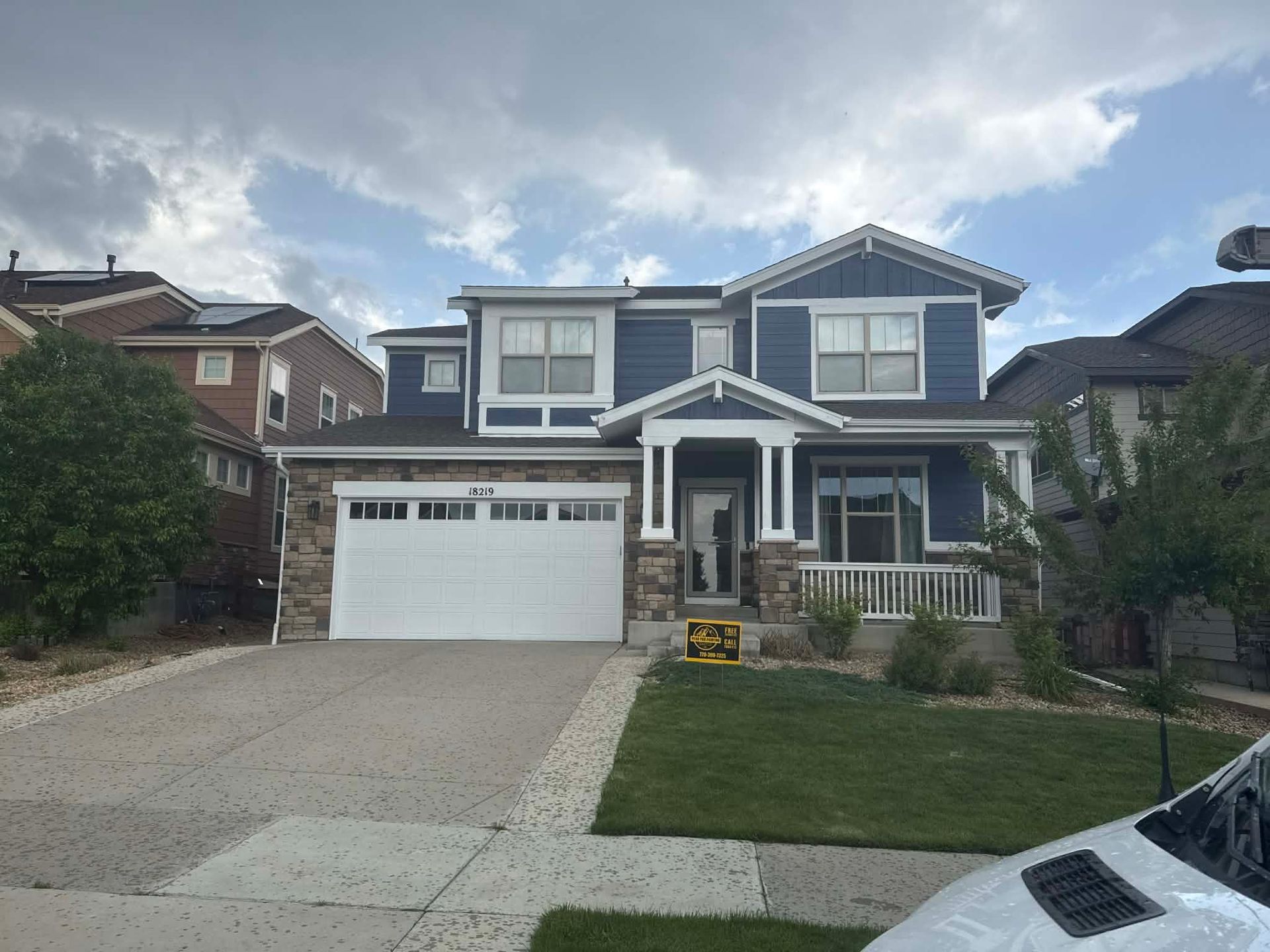 A two-story house with dark blue siding, stone veneer accents, a white garage door, and a small front porch under a sky.