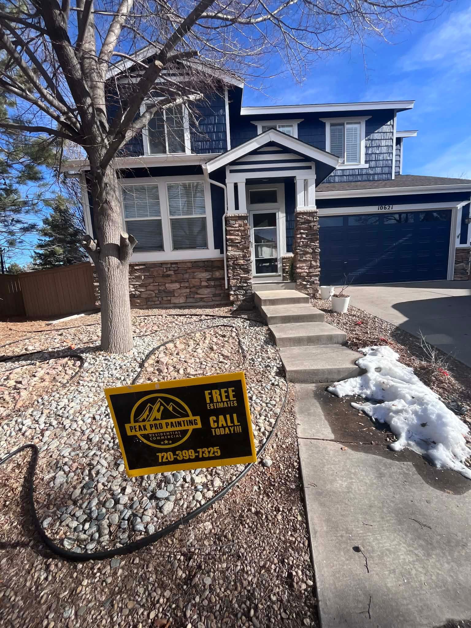 A blue, two-story house with a stone-accented porch, a rock garden, and a lawn sign in the front yard.