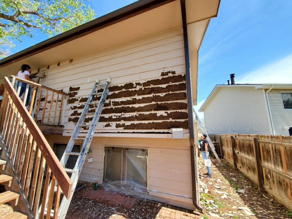 Two people stand on ladders repairing the light-colored wooden siding on the exterior of a house.