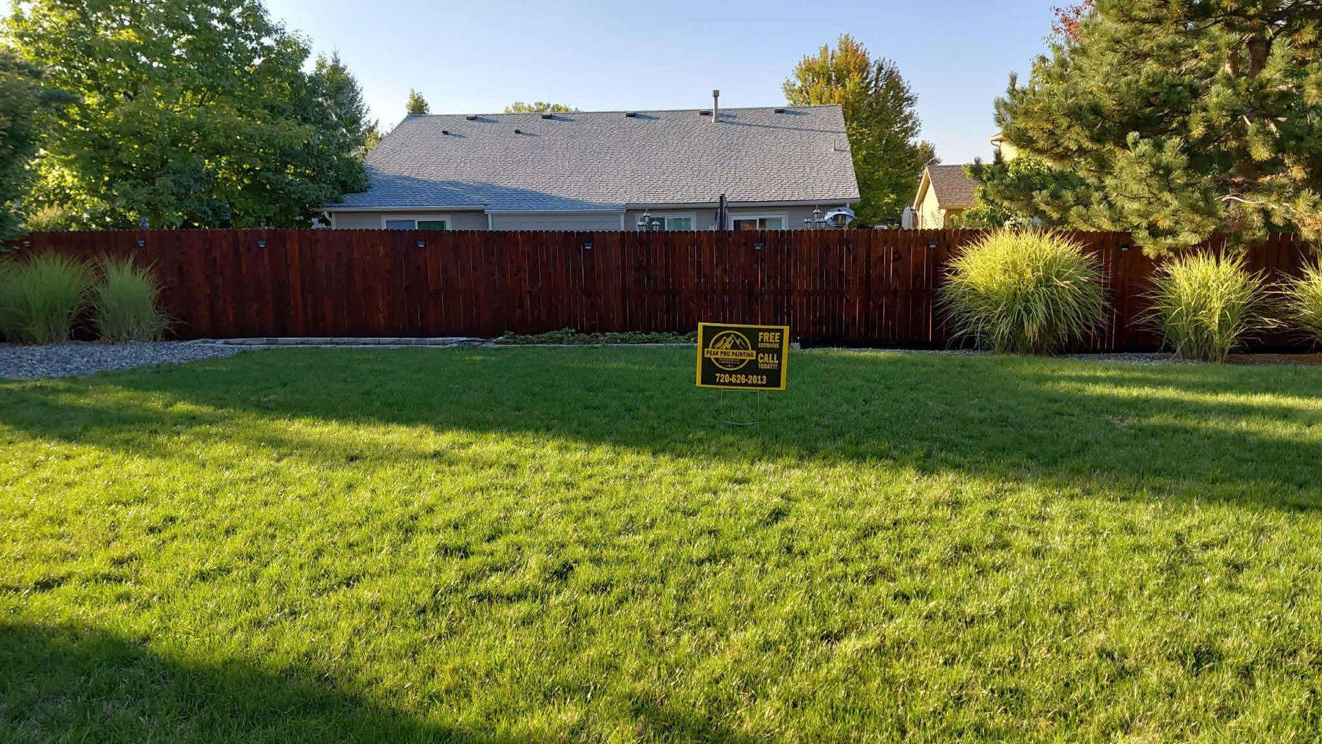 A small sign sits in the center of a grassy backyard in front of a reddish-brown wooden fence and a house.
