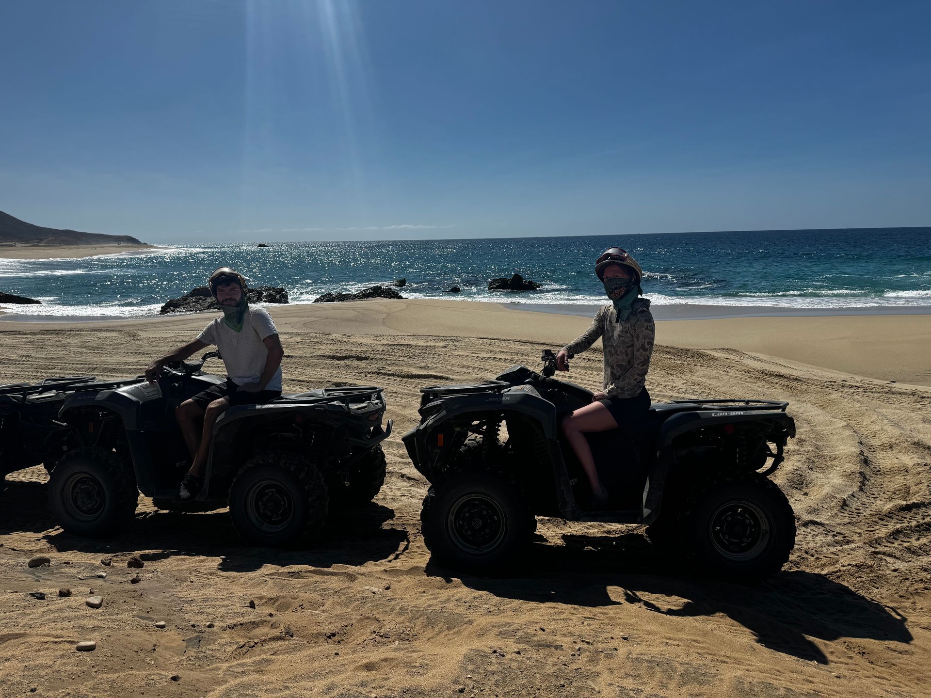 Two people sit on ATVs parked on a sunny, sandy beach by the ocean.