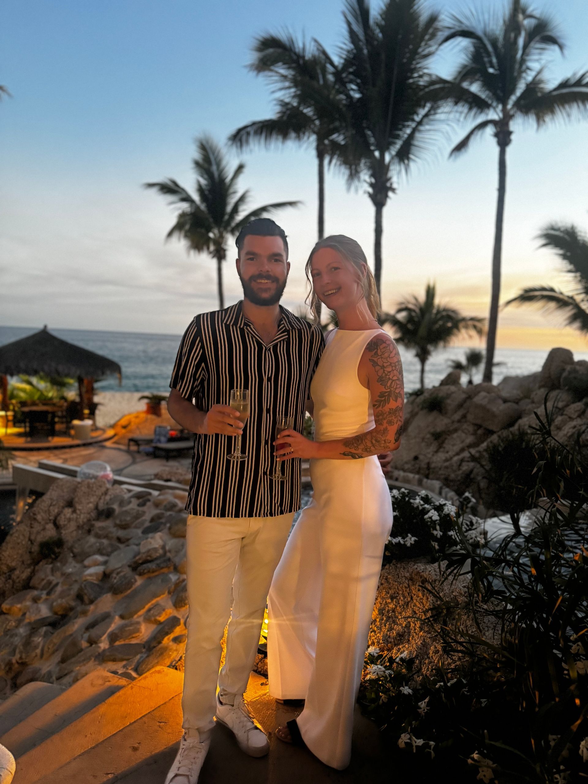 A couple stands together holding glasses at sunset on a beach, with palm trees and a resort in the background.