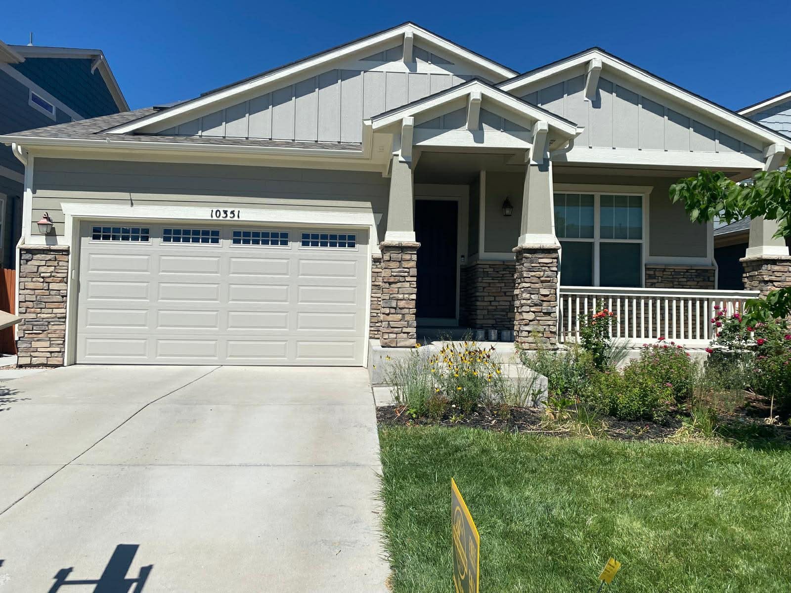 A suburban single-story house with a white garage, gray siding, stone accents, and a front porch under a blue sky.