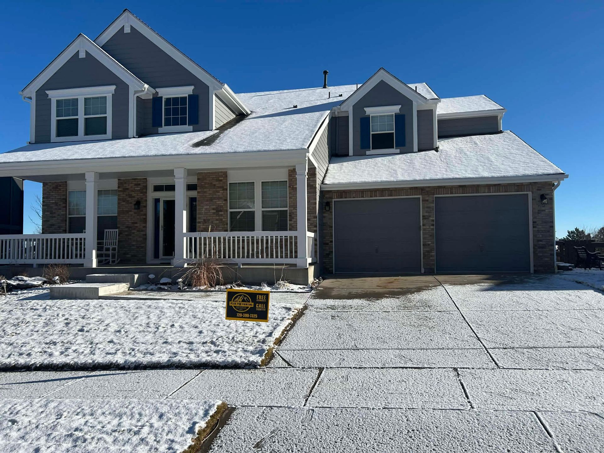A two-story grey and stone house with a snowy yard, porch, and two-car garage under a clear blue sky.