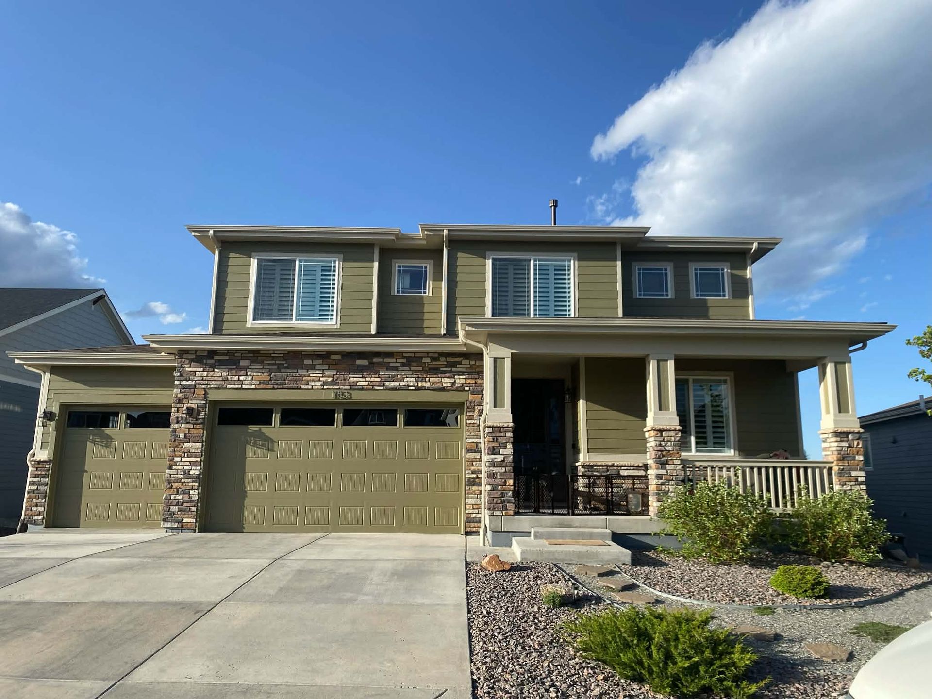 A two-story olive green suburban house with a stone-accented facade, a three-car garage, and a front porch.