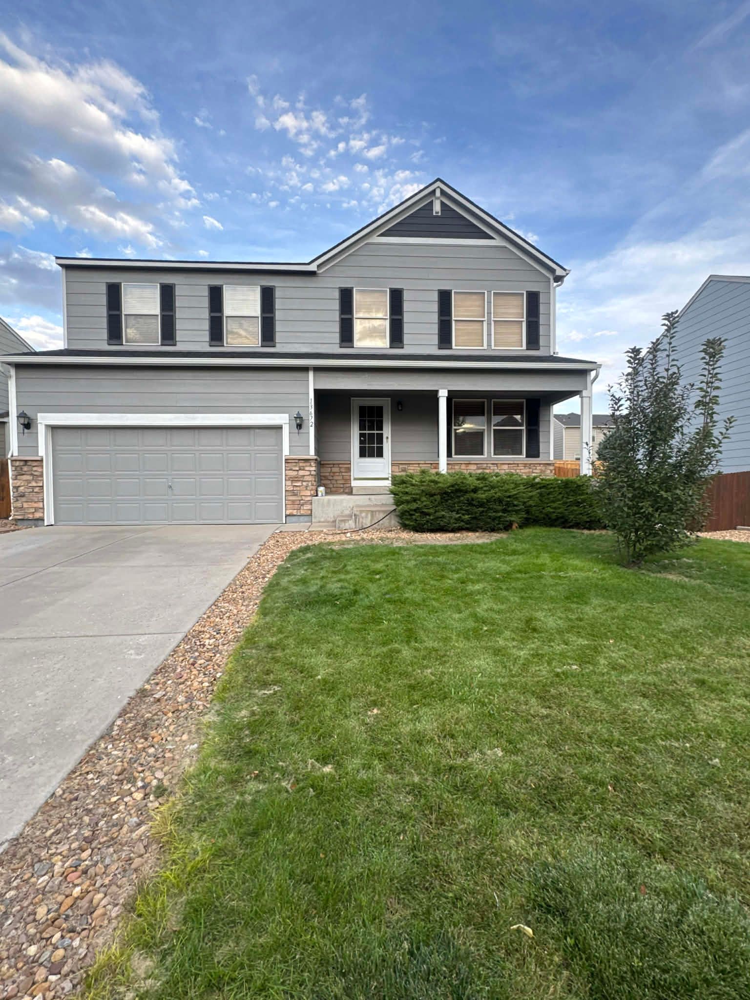 Two-story, gray house with a front-facing garage, tan stone accents, and a grass lawn under a blue sky.