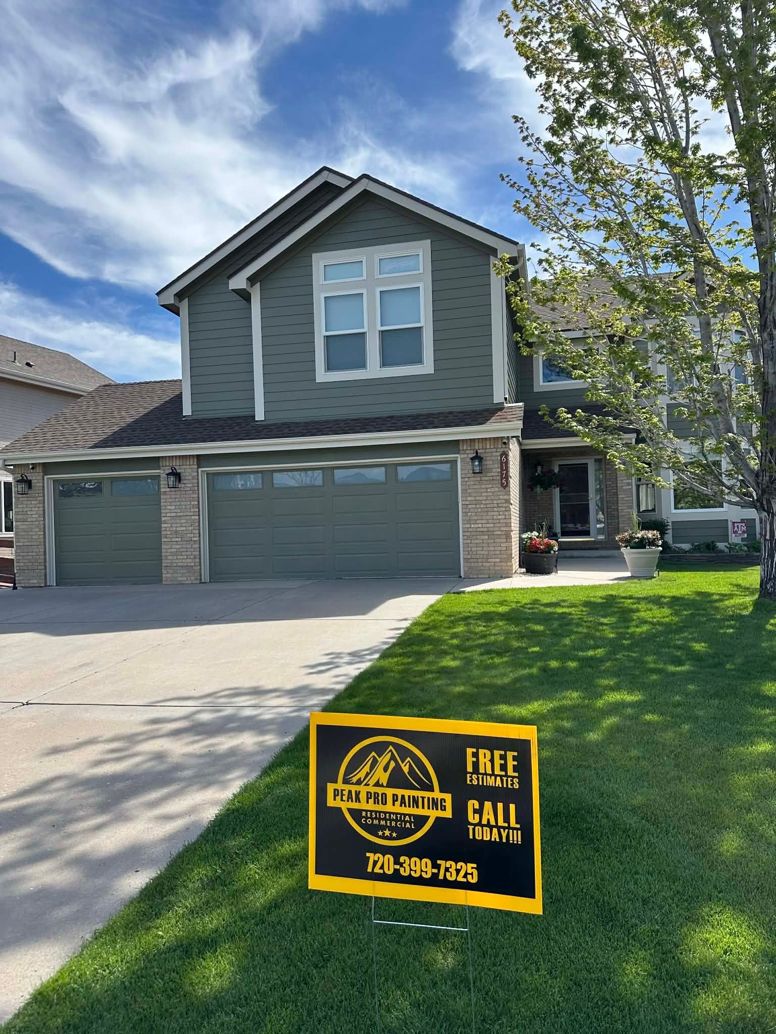 A two-story green suburban house with a three-car garage, front lawn, and a marketing sign in the yard.