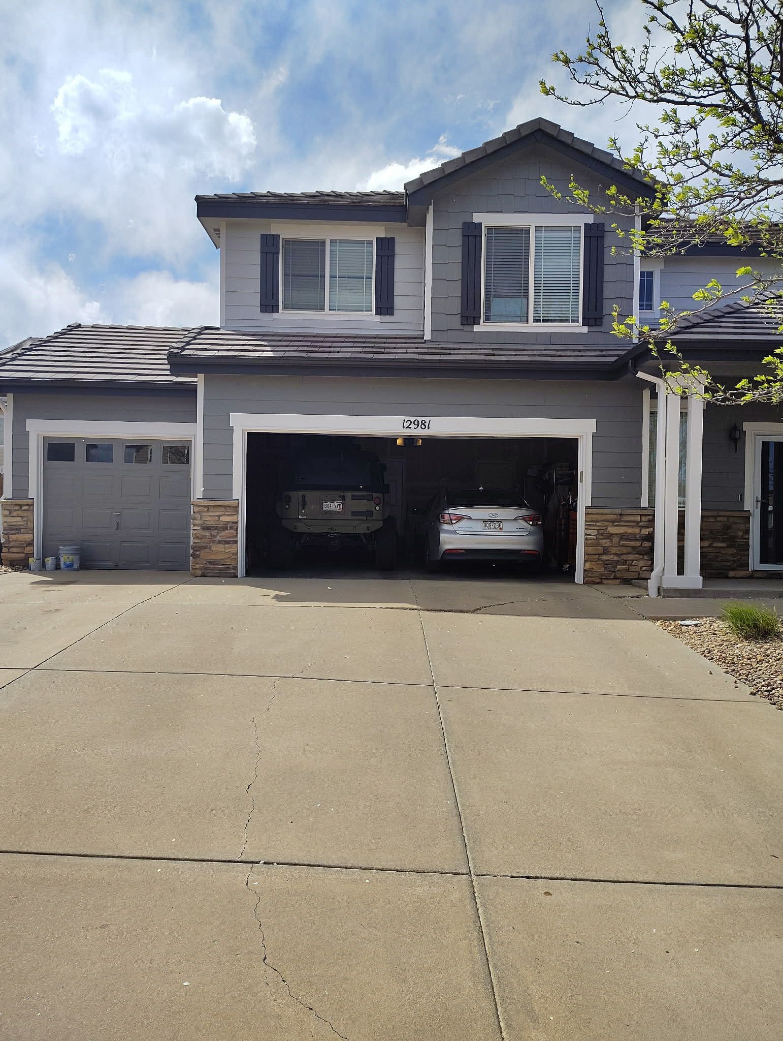 A two-story grey residential house with a multi-car garage, a concrete driveway, and some visible fire damage on the roof.