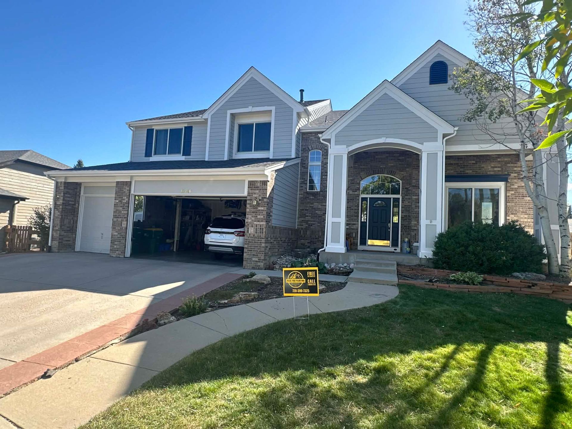 A two-story suburban home with stone and light gray siding, a garage, a front porch, and a lawn under a blue sky.