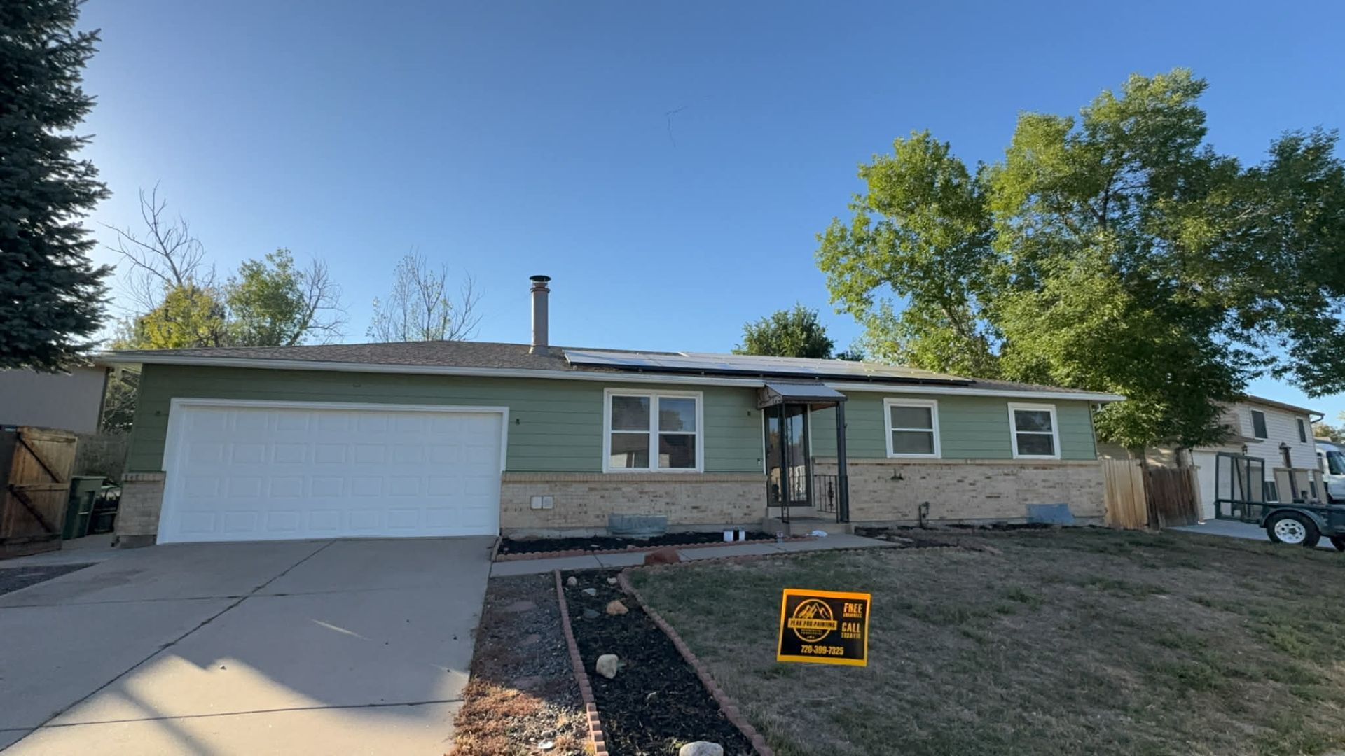 A one-story suburban home with a green horizontal siding, stone base, white garage, and a yellow sign on the front lawn.