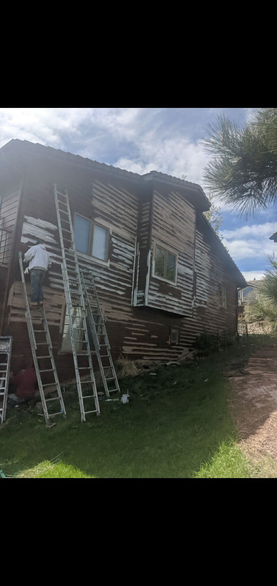 A worker on a tall ladder scrapes old, peeling paint off the wooden siding of a two-story house on a sunny day.