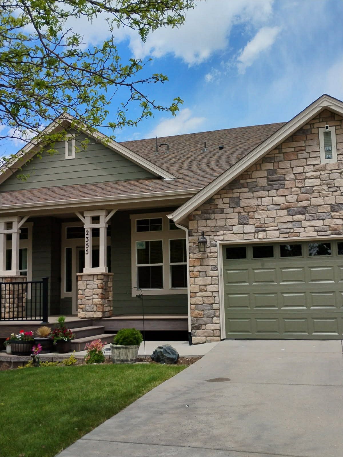 A single-story craftsman home with olive green siding, a stone facade, a front porch, and an attached two-car garage.