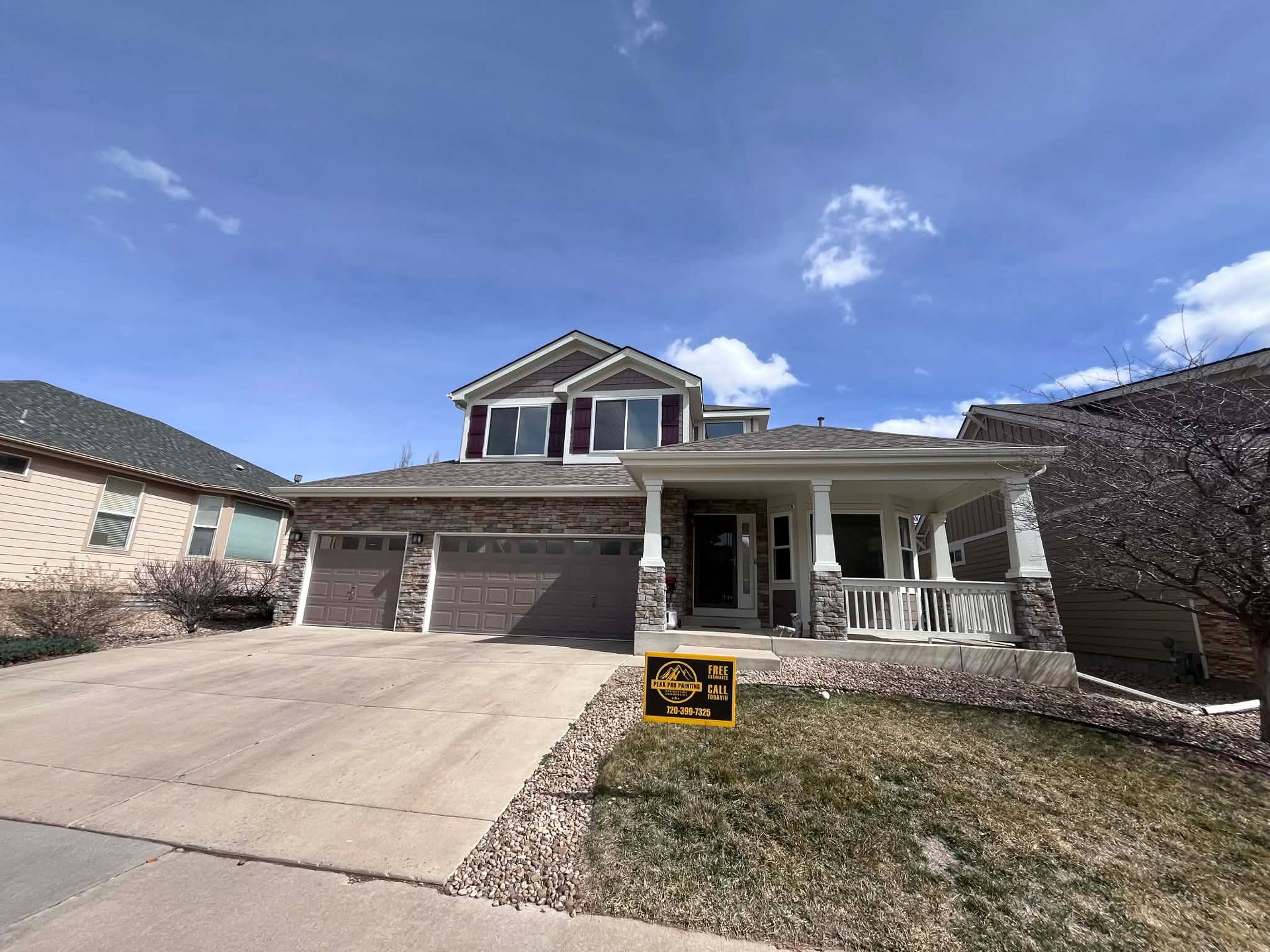 A two-story suburban home with a stone and tan facade, a two-car garage, and a covered front porch under a sunny sky.