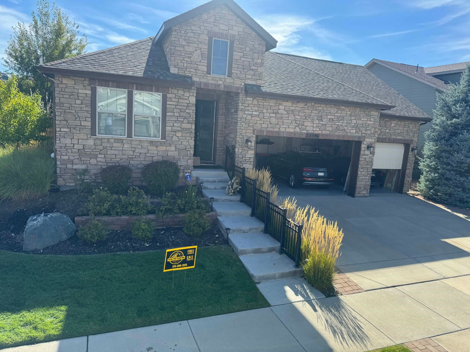 A tan stone house with a multi-level roof, a front porch, and a driveway with a parked car on a sunny day.