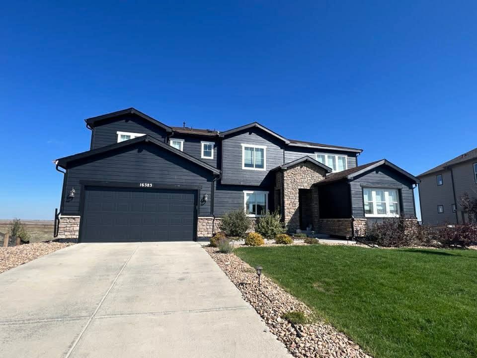 A two-story dark blue house with a stone-accented facade, a two-car garage, and a manicured lawn under a clear blue sky.